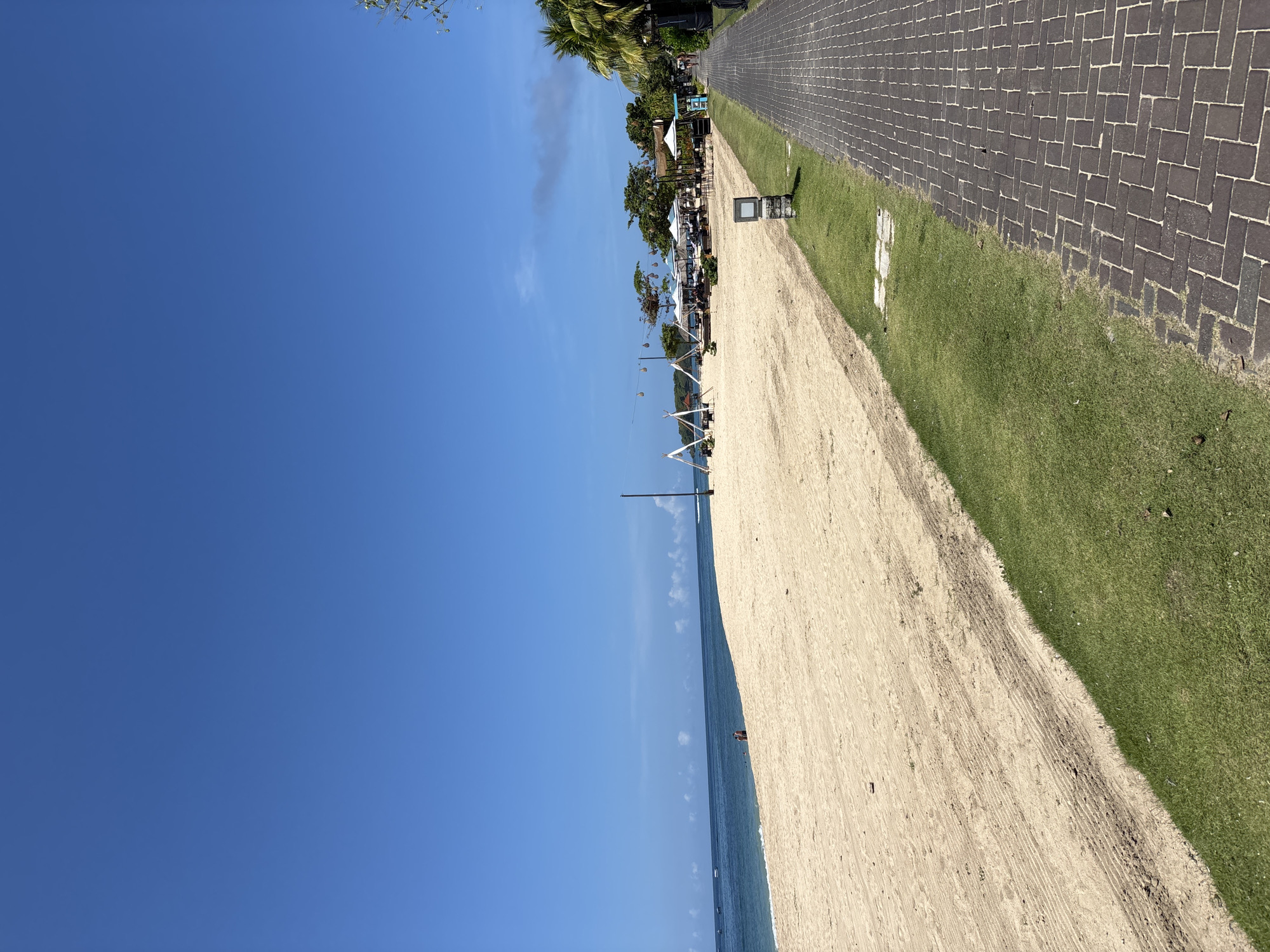 The image depicts a beach scene in Kuta Selatan, Indonesia. The sky is a clear blue. The beach stretches from the bottom right to the center of the image, showing light-colored sand with visible tire tracks. The ocean is a deep blue, with small boats visible.
A strip of green grass runs parallel to the beach, separating it from a brick-paved walkway. The walkway is composed of dark gray rectangular bricks arranged in a herringbone pattern.
Along the edge of the walkway, closer to the beach, are several beachside establishments with white canopies and various seating arrangements. Trees and other vegetation are scattered among these establishments.