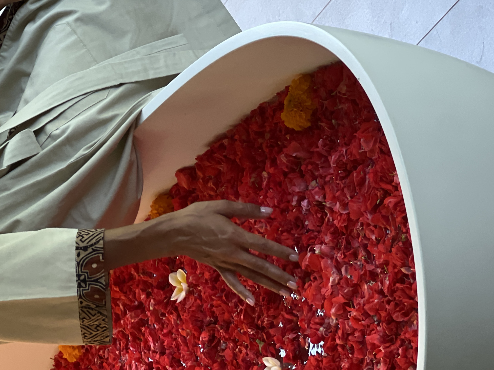 In Ubud District, Indonesia, a person's hand with manicured nails is submerged in a white bathtub filled with red flower petals and a few yellow flowers. The person is wearing a light-colored robe with a patterned trim on the sleeve. The bathtub is partially visible and appears to be made of a smooth, matte material. The environment suggests a spa or a similar setting designed for relaxation and wellness.