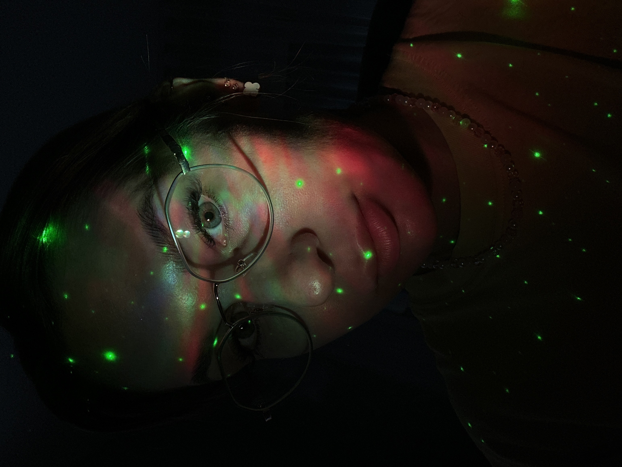 A close-up portrait shows a person lying on their side, illuminated by green and red laser lights. The individual is wearing round glasses and a light-colored beaded necklace. Light scatters across the skin of their face and neck, as well as their shirt. Details such as earrings, hair, and facial features are visible, though distorted by the light effects. The environment is dark. No specific landmarks indicate that the image was taken in Chaikovsky, Russia.
