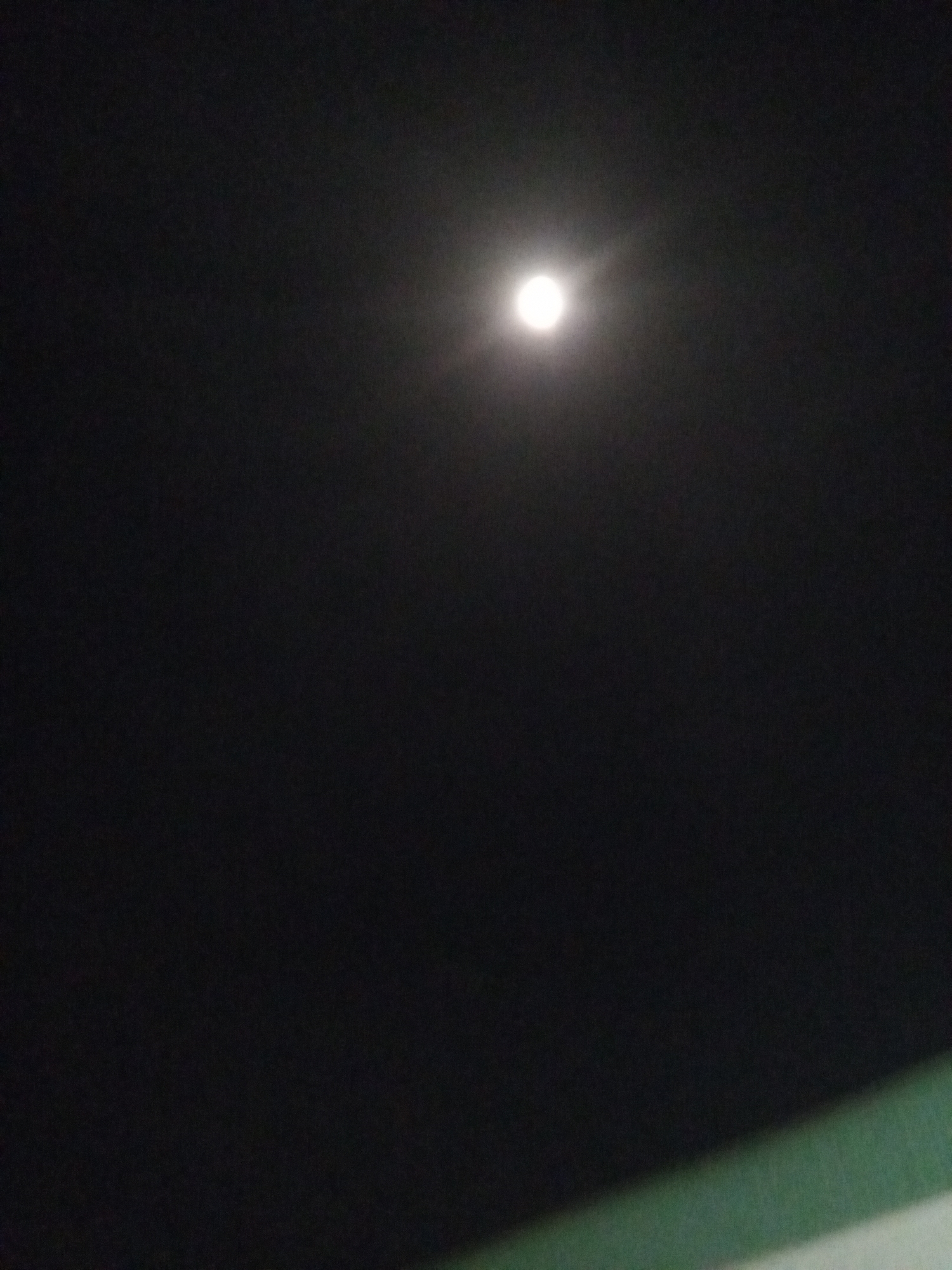 The image captures a clear night sky, dominated by a bright, circular celestial object, identified as the moon. The moon is prominently positioned slightly above the center of the frame and emits a strong, white light, surrounded by a soft, diffused halo against the dark background. This halo suggests either slight atmospheric haze or the effect of the camera's lens capturing the intense light. The vast majority of the frame consists of a deep, unblemished black, characteristic of a cloudless night sky. In the bottom right corner, a slanting, blurred element with a gradient of green fading into white is partially visible, likely indicating the top or edge of a nearby structure or foreground object. No people or specific activities are depicted. The time of day is clearly night. No text is visible in the image. While the location context of Jos, Nigeria is provided, there are no specific visual cues within the image itself to confirm this geographic location.