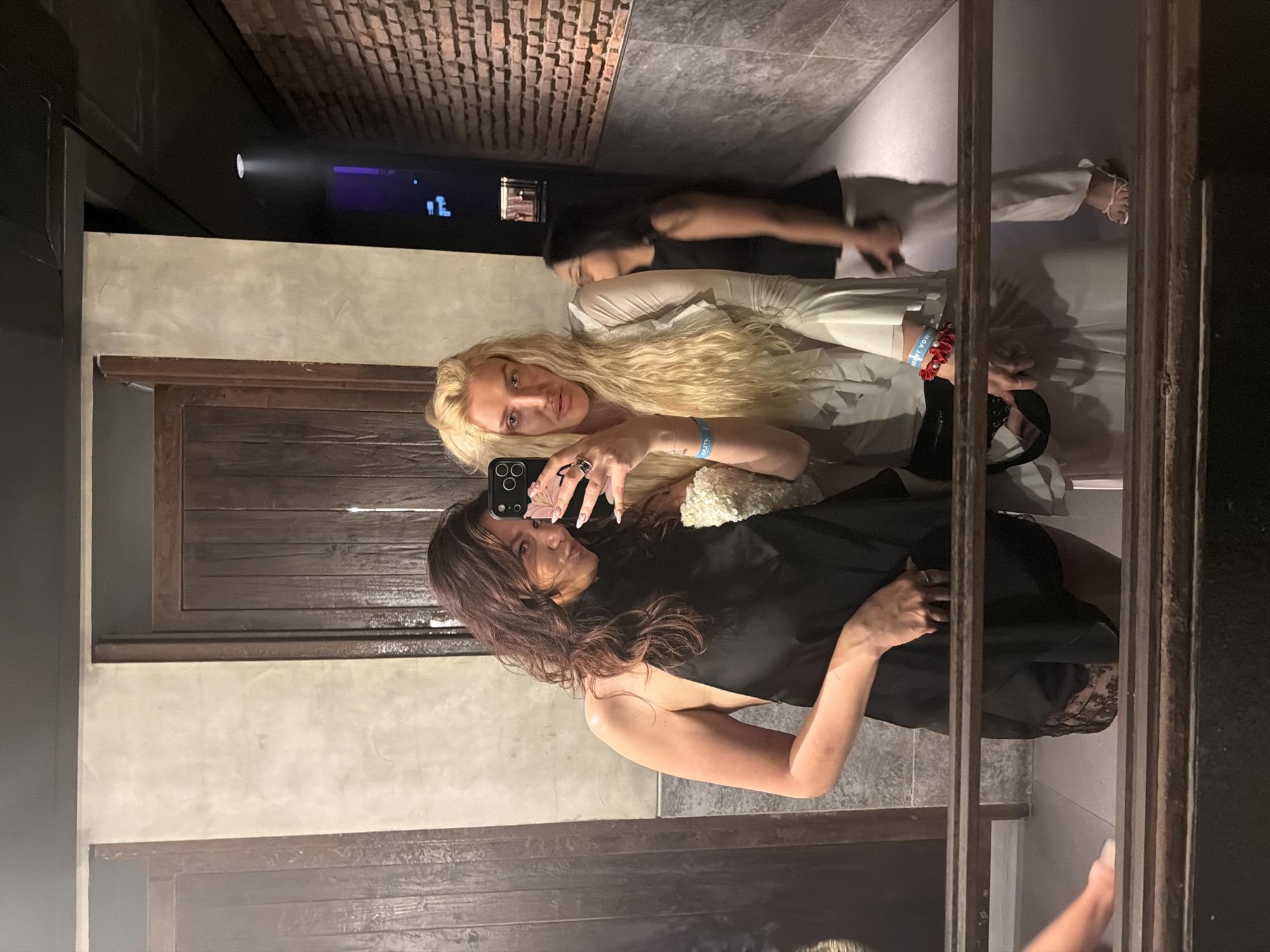The image, oriented upright, depicts two women taking a mirror selfie in a dimly lit indoor setting. The woman on the left, with long dark brown hair, wears a black sleeveless top and looks directly at the camera with a subtle expression. The woman on the right, with long blonde hair, holds a black smartphone with a patterned case to capture the reflection. She wears a light-colored, possibly white, long-sleeved garment. Her left wrist displays a blue bracelet with partial text, "YOUTH" and "BALI", and a red beaded bracelet. Rings adorn her fingers.
The background shows a mix of architectural elements. Dark wooden panels, possibly doors or wall features, are visible behind the women. Above them, in the mirror's reflection, is a ceiling or upper wall constructed from exposed reddish-brown bricks. Sections of light grey concrete or stucco walls are also present. A narrow, dark metal frame runs vertically in the foreground, part of the mirror's structure or a decorative divider.
Further in the reflected background, a third person with dark hair, wearing a dark top and light pants, is partially visible, appearing to walk away. Artificial lighting creates the ambiance; a bright circular light source projects a beam, and on a distant dark surface, blue pixelated figures resembling people are displayed. The overall scene suggests an evening event or a stylish indoor venue, consistent with the provided location context of North Kuta, Indonesia.