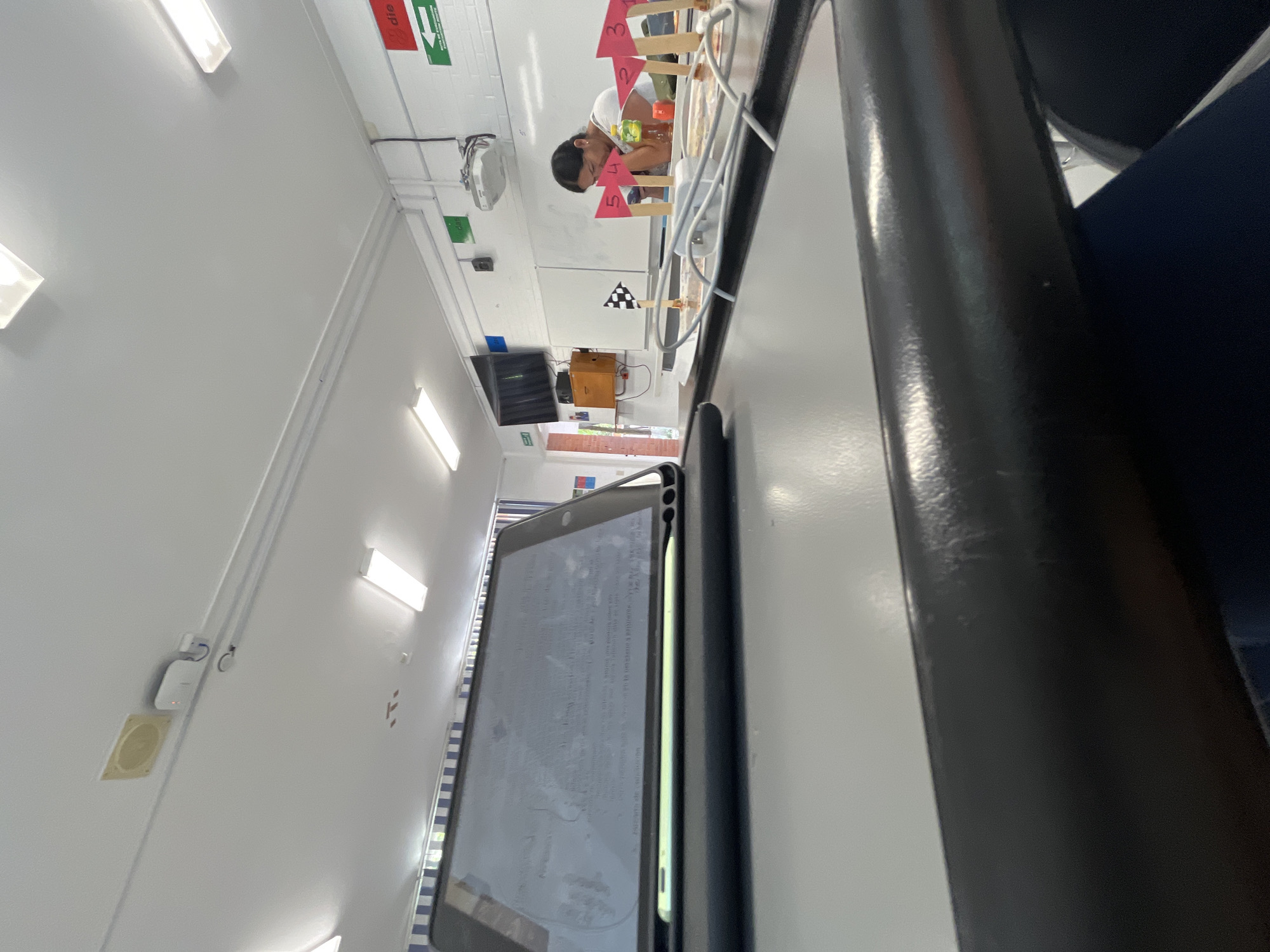 The scene appears to be indoors, possibly a classroom or laboratory setting. A young person is working on an experiment or project with numbered markers and a checkered flag. The location is within the city of Puebla, Mexico, given the "die" sign written in German. The environment is clean and well-lit with fluorescent lighting and white walls. There is a tablet, possibly an iPad, close to the camera, displaying text on the screen. A black chair is also in the frame. The focus seems to be on the person working on the project and the overall environment.