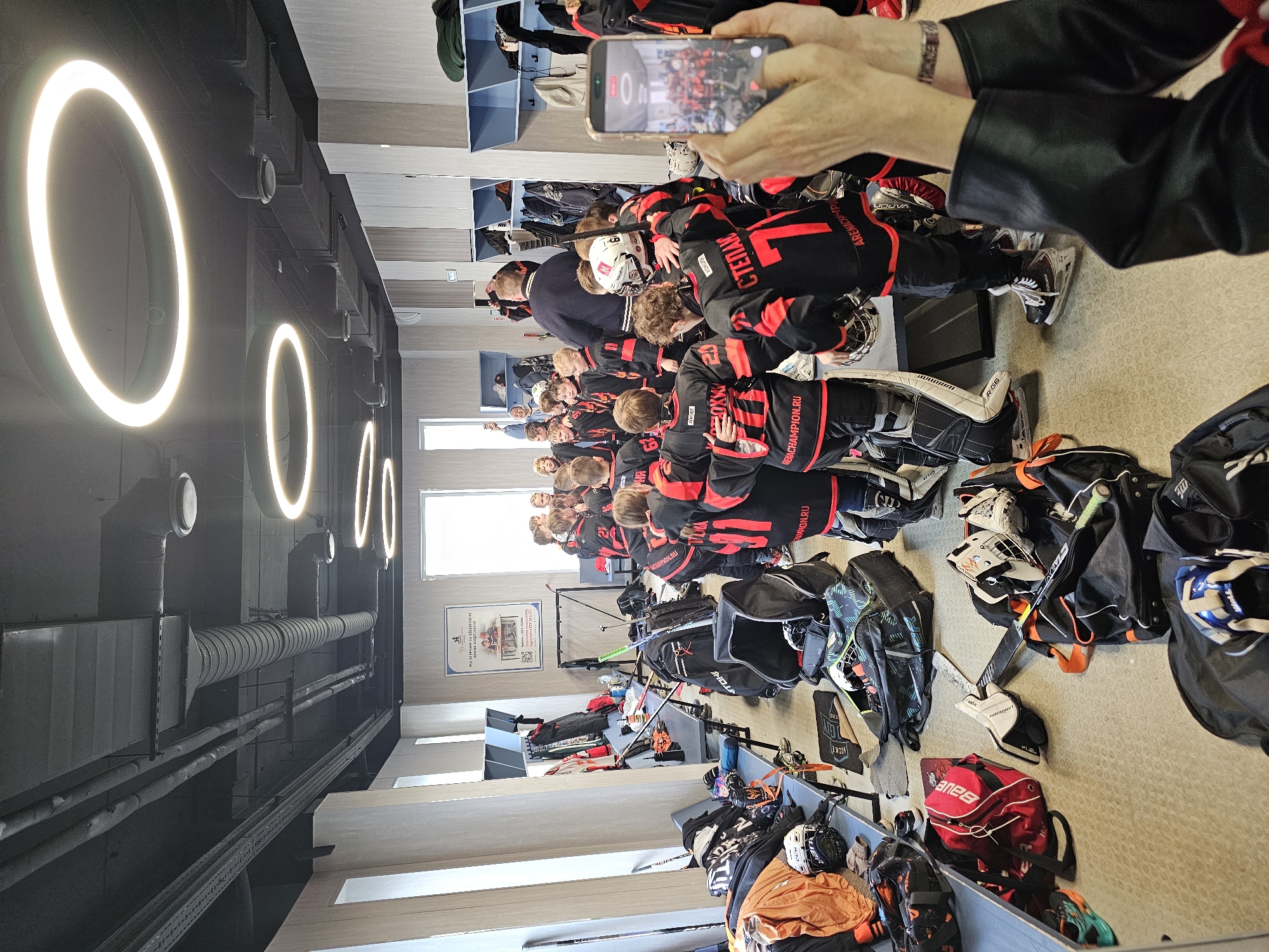 Here's a detailed description of the image:
**Scene:** A group of young hockey players in black and red jerseys are huddled together in what appears to be a locker room.
**People:**
* A team of youth hockey players (approximately 20) wearing matching jerseys. Several jersey names and numbers are visible including "Степан" (Stepan) and the number "7".
* An adult male, possibly a coach, stands behind the group.
* Another adult is taking a photo or video of the team with a mobile phone.
**Objects:**
* Hockey equipment: Helmets, shoulder pads, sticks, gloves, and bags are scattered around. Goalie equipment is visible as well.
* Lockers: Light blue lockers are built into the walls.
* Ceiling: the ceiling has round, white lights.
* Wall: there is a poster on the wall in the middle of the room, and some windows.
**Actions/Interactions:**
* The hockey players are huddled together, likely for a pep talk or team moment.
* One person is using a mobile phone to capture the moment.
**Environment:**
* The scene appears to be a locker room.
* Based on the jersey and helmet details, and the presence of Russian text ("ACHAMPION.RU," "ARENACHIMPI"), the team is likely from Russia.