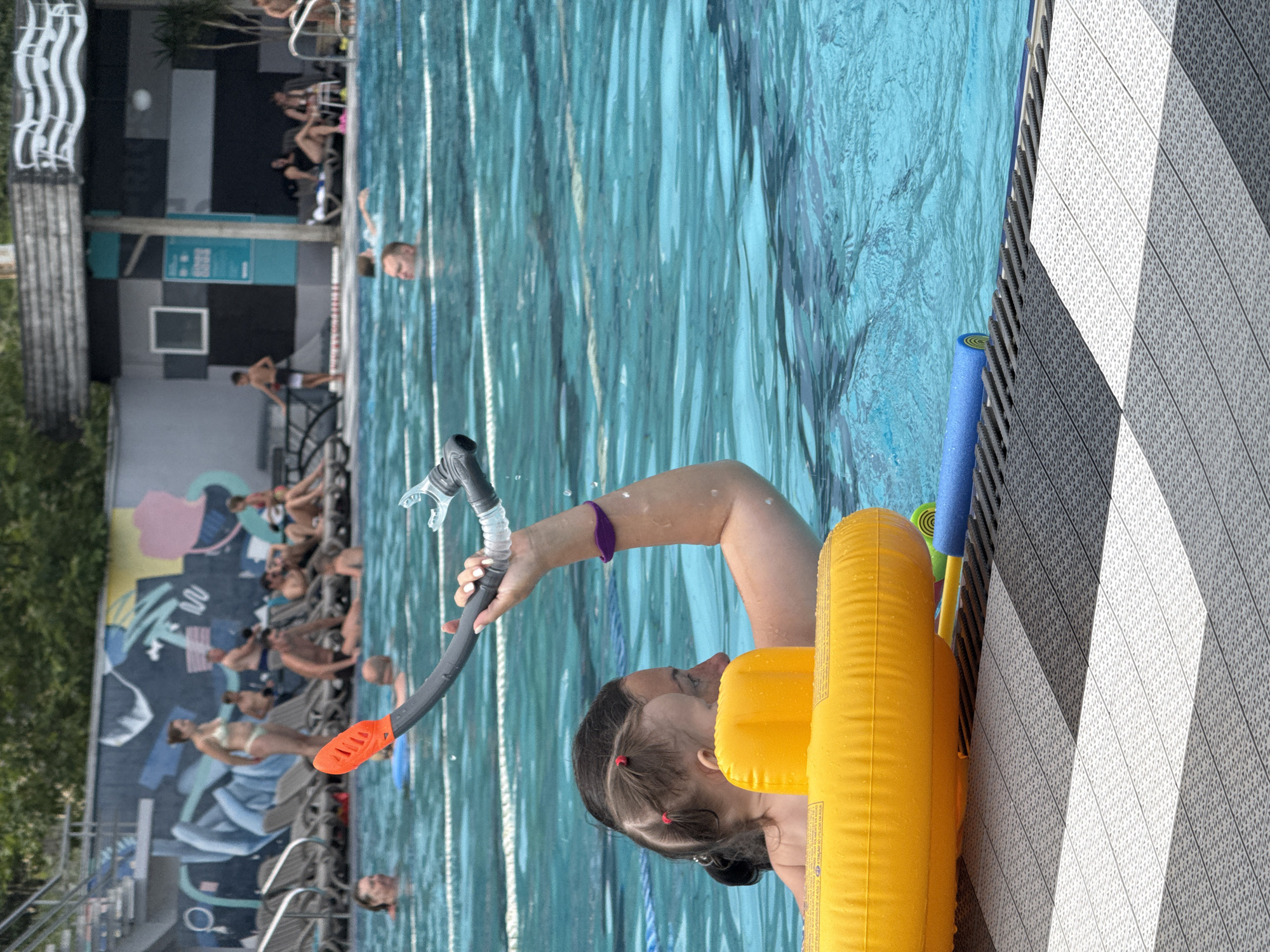 In the foreground, a woman with pigtails and a purple bracelet is in a swimming pool, holding an orange snorkel and mask. A yellow inflatable ring is around her. In the background, other people are visible in the pool and on lounge chairs next to the pool. The pool area has lounge chairs, a mural on a wall, and buildings in the distance. The image appears to be taken at a public pool on a sunny day. The location is identified as Yalta, Ukraine.