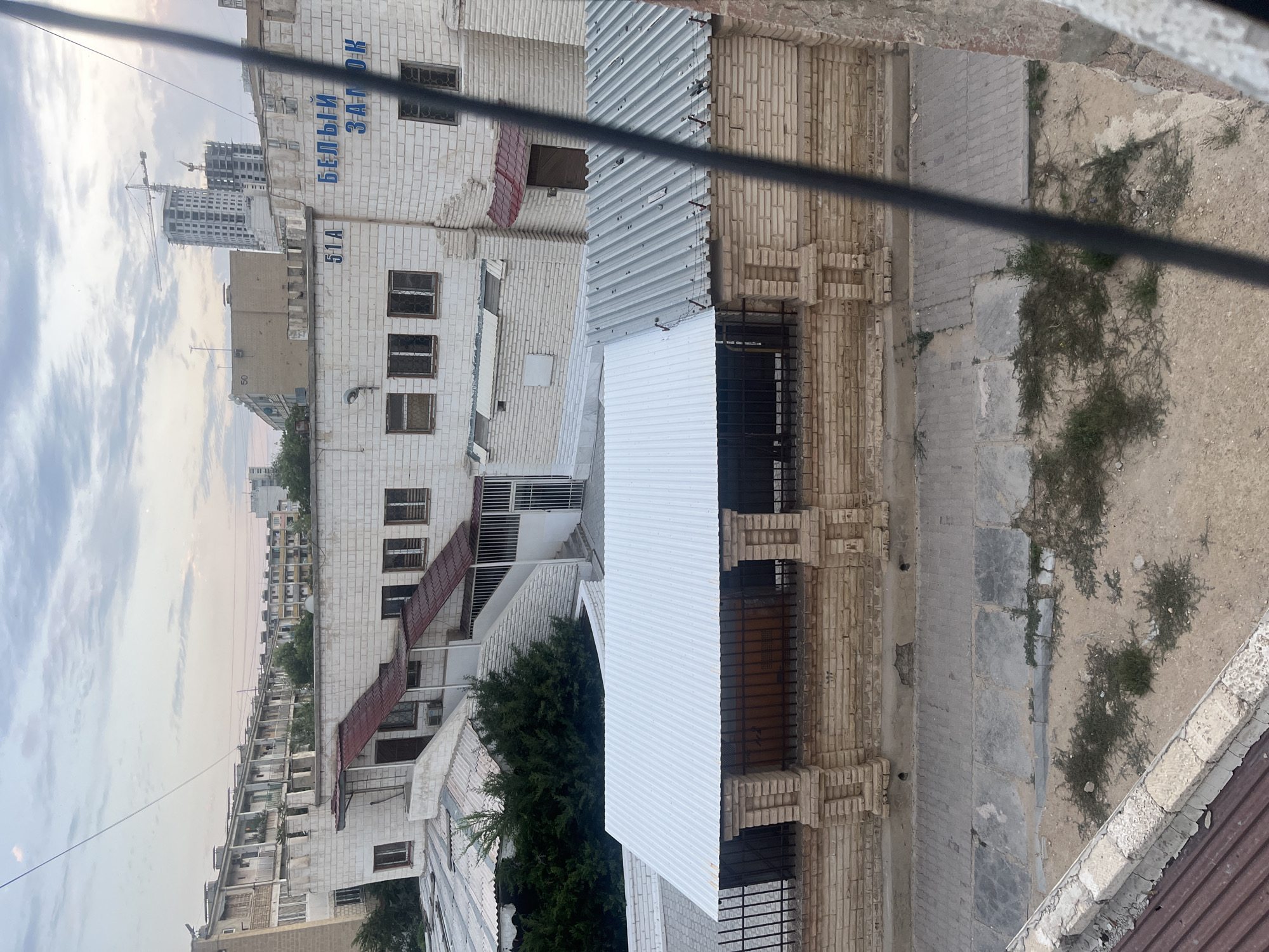 This is a rotated, medium-angle shot of an urban scene in Aktau, Kazakhstan, focusing on a multi-story building. The building's facade is constructed from white brick, with a section featuring exposed beige brickwork and decorative brick columns. Several windows are visible, some with metal grates. A prominent feature is a white, corrugated metal roof extending over part of the structure, with a section of a darker, possibly red, corrugated metal roof above it. Adjacent to the building is a sidewalk with a brick pattern, bordered by a patch of dry, sandy ground with sparse green vegetation. In the background, more residential buildings and construction sites with cranes are visible, suggesting ongoing development. The sky above is cloudy with a soft, diffused light, indicating either early morning or late afternoon. The overall atmosphere is one of an ordinary urban environment. The text "БЕЛЫЙ ЗАЛОК" and "51А" is visible on the white brick facade.