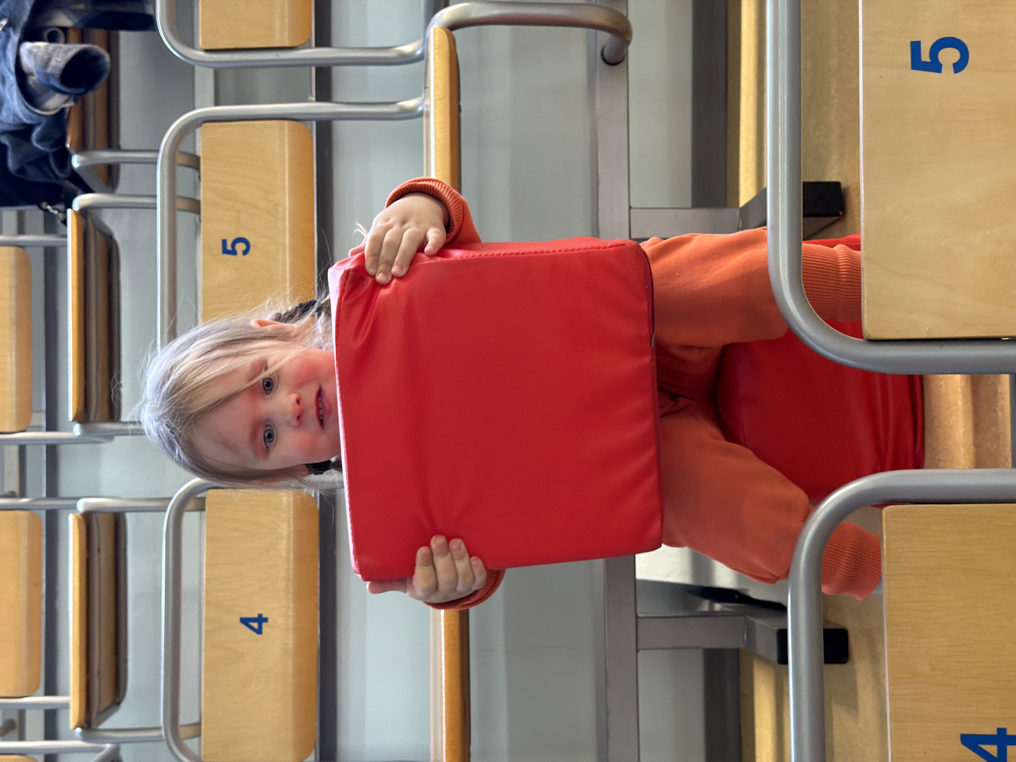 A child in an orange sweater is holding a red, square cushion while sitting in a classroom in Chaikovsky, Russia. The child is blonde with blue eyes and is facing the camera. The classroom contains rows of chairs with metal frames and wooden seats, numbered 4 and 5. There is a jacket on the top row of the chairs.