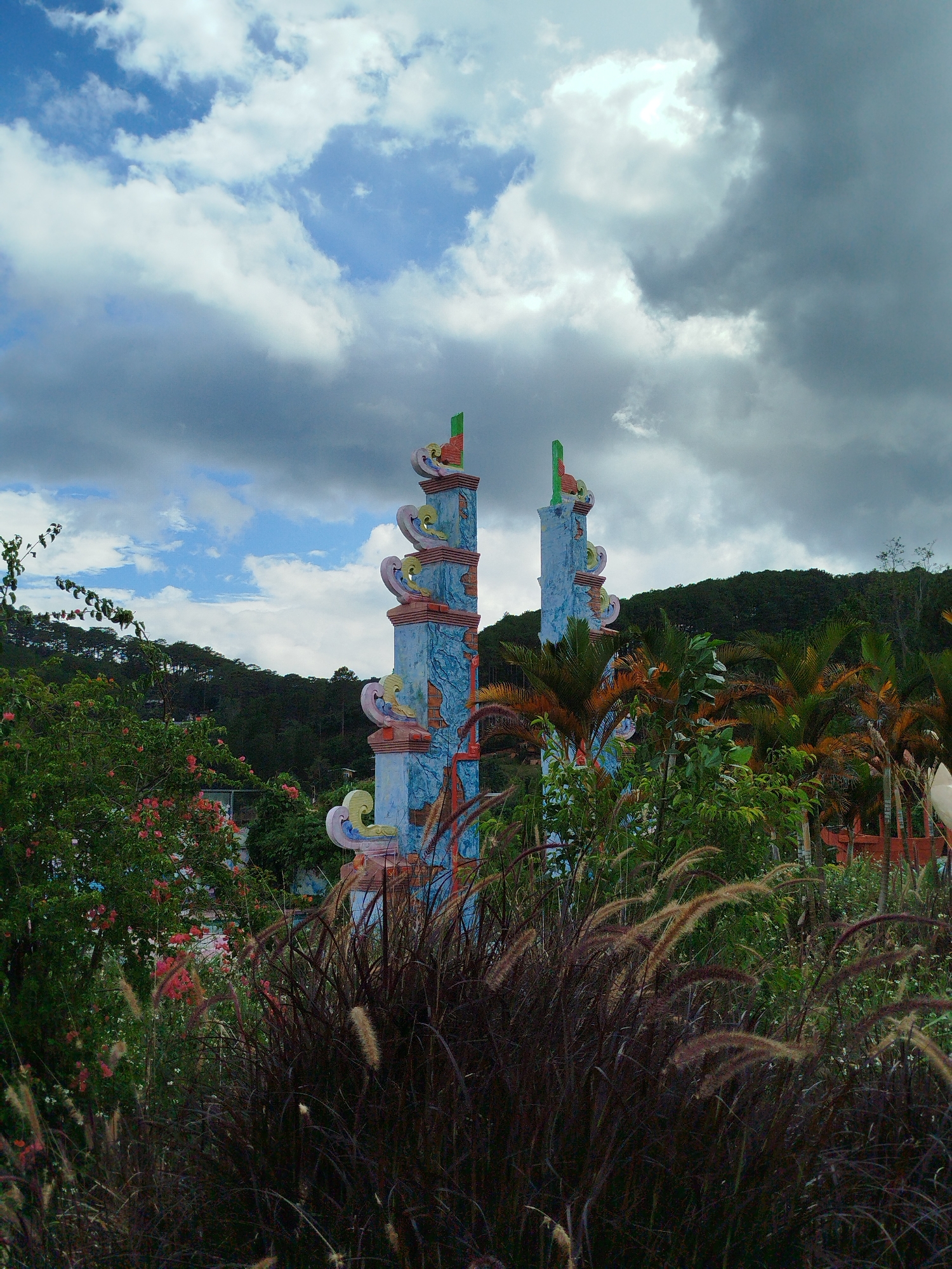Two elaborately decorated, light blue-colored towers with orange, green, pink, and white embellishments stand partially obscured by dense vegetation in a daytime shot. Behind the towers, a landscape of green trees and vegetation rises into a distant hill or mountain range. The sky is a mix of dark, ominous clouds and patches of brighter, light blue sky with white clouds. In the foreground, there are thick bushes and grasses. This scene is suggestive of a public space in the city of Da Lat, Vietnam.