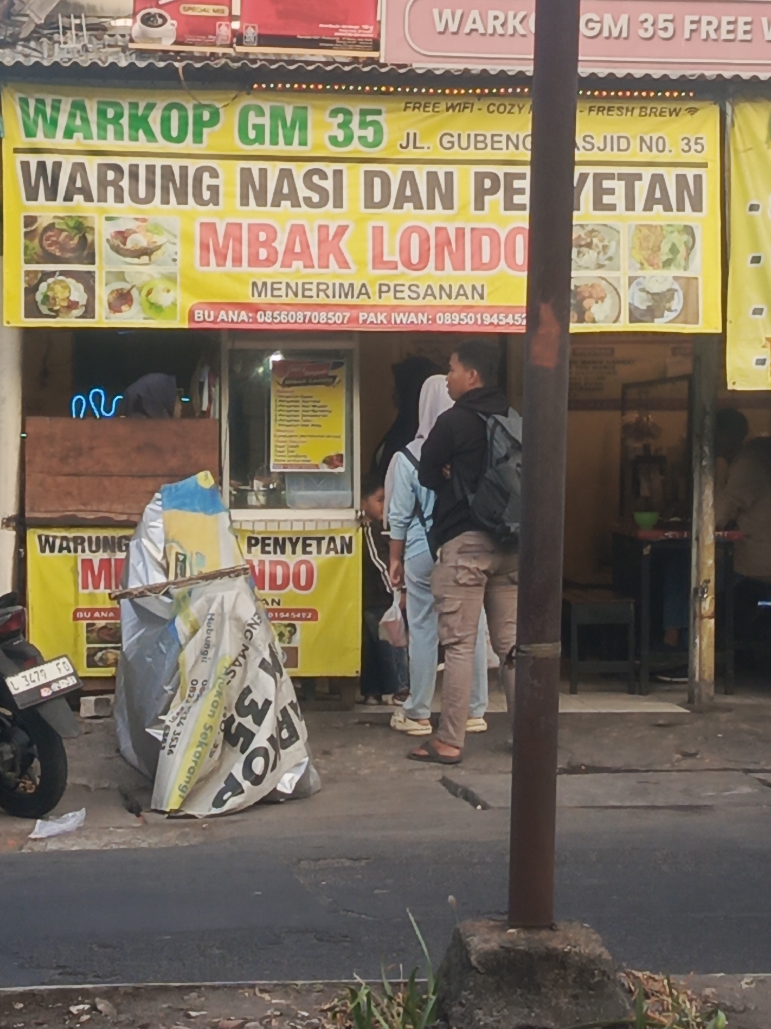 This is a street-level, medium-shot, outdoor photograph taken in Surabaya, Indonesia, likely during the daytime. The image is dominated by the facade of a food establishment. A prominent yellow banner across the front reads "WARKOP GM 35 JL. GUBENG MASJID NO. 35". Below this, another banner announces "WARUNG NASI DAN PERYETAN MBAK LONDO". Several images of food are displayed on both banners. To the left, a motorcycle is partially visible, with a license plate reading "L 3479 FD". A large, white, tarp-covered object, possibly a temporary stall or display, is situated in front of the establishment. Several people are present; one man in a black hoodie and backpack stands with his arms crossed, facing away from the camera, while others stand behind him. Some are looking towards the entrance of the establishment. The background shows the interior of the warung with tables and chairs, and other patrons can be seen seated. The environment appears to be a busy street, with a sidewalk and asphalt road in the foreground.