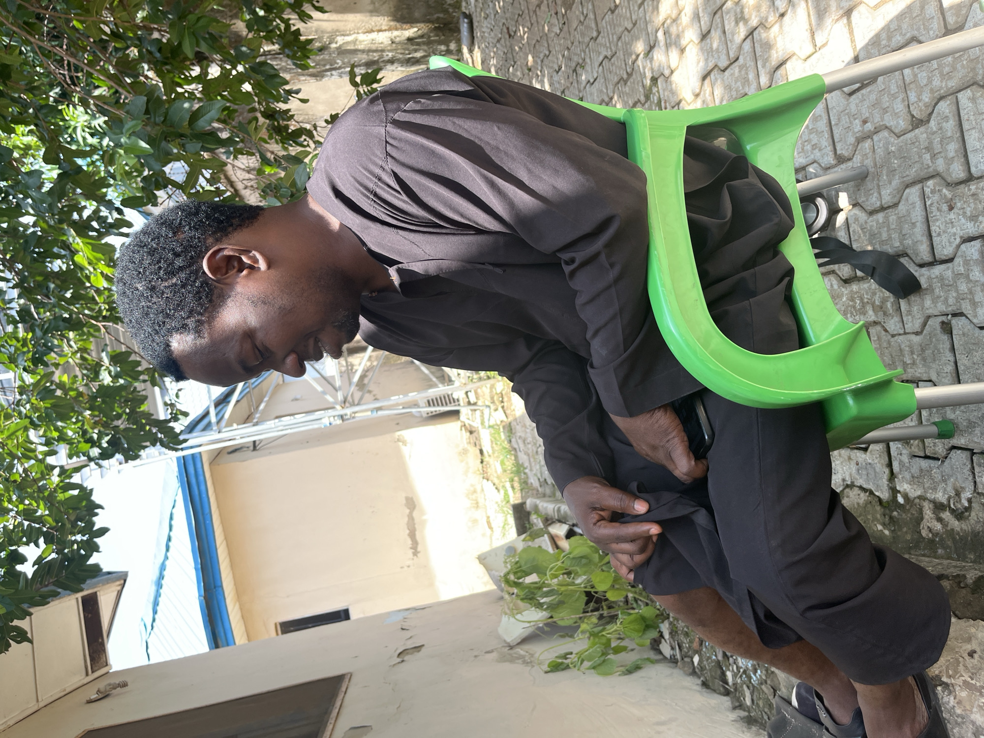 A man sits on a bright green plastic chair in an outdoor setting. He is wearing a black traditional outfit. The man is facing to his right and looking down. He appears to be resting his hands on his lap. A black mobile phone is placed on his right thigh. The ground beneath him is paved with interlocking bricks, and there are trees and a building visible in the background. The overall scene suggests a relaxed, casual atmosphere.