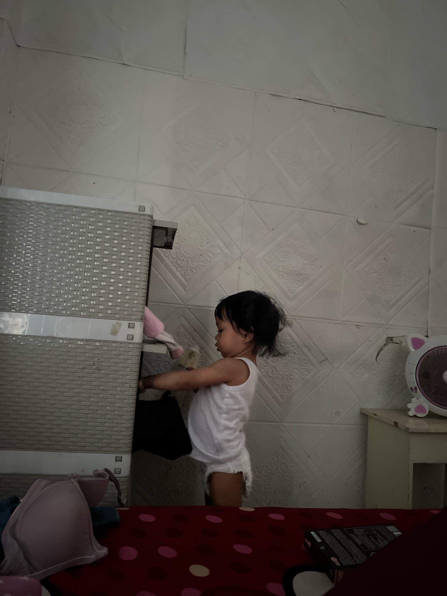A young child wearing a white tank top and diaper is standing next to a grey plastic storage cabinet. The child's hands are reaching into one of the cabinet's drawers. A pink object, possibly a stuffed toy, is visible sticking out of the same drawer. The background is a white tiled wall with a diamond pattern. To the right of the child, a small white table holds a fan with a cat-like design. The foreground features a red surface with pink polka dots, possibly a bed or blanket, with a pink bra lying on it. The scene appears to be indoors, in a domestic setting. The lighting suggests it could be daytime. No visible text is present in the image.
