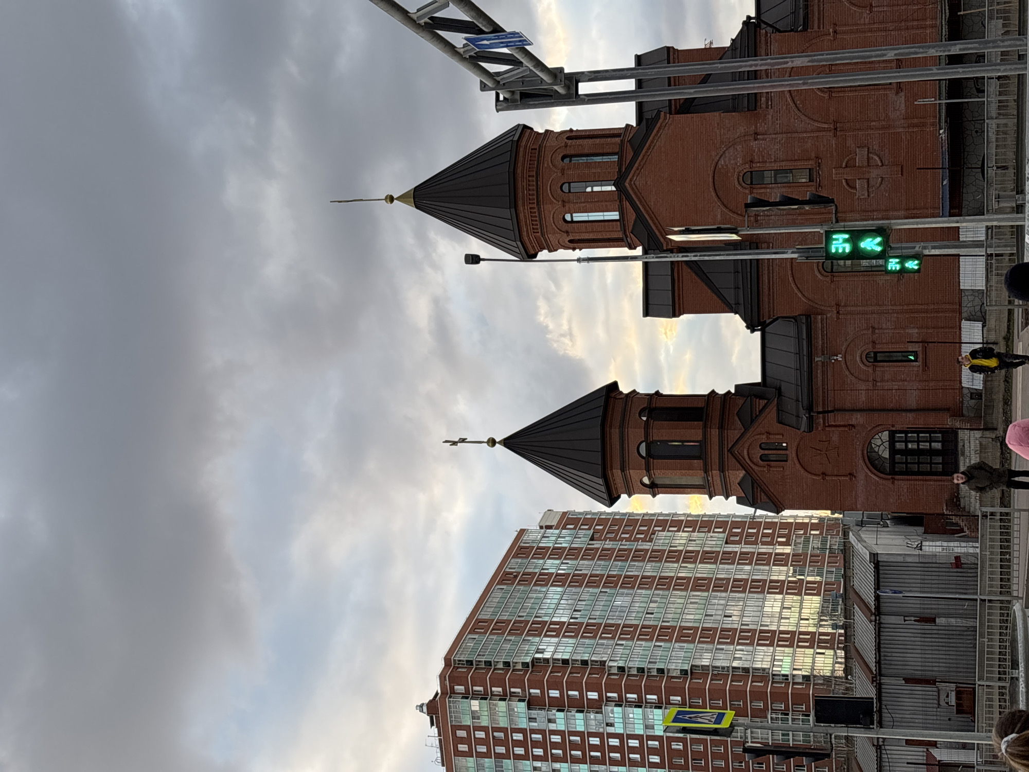 The image captures an urban street scene in Perm, Russia, under an overcast sky with patches of brighter light suggesting late afternoon or a cloudy day. The perspective looks up at two prominent buildings and street infrastructure.
The most striking subject is a large building with reddish-brown brickwork, featuring two distinct towers topped with dark, conical roofs and slender crosses, characteristic of an Armenian Apostolic Church. The church's facade displays arched windows and decorative brick patterns, including a circular cross design.
Adjacent to the church, a tall, modern residential or office building rises, also constructed with reddish-brown brick and numerous large, reflective windows that mirror the sky.
In the foreground and mid-ground, street elements are visible, including metal overhead structures supporting traffic lights. One prominent set of traffic lights shows green pedestrian signals: a walking person and a left-pointing arrow. Another smaller green arrow is visible below it. A blue street sign with a white arrow, likely indicating a turn or one-way street, is attached to the overhead structure. A yellow diamond-shaped crosswalk sign is also visible near the modern building. Several people are on the sidewalk, including one person in a yellow jacket and another in a dark jacket near the church entrance. A corrugated metal barrier or fence runs along the sidewalk.