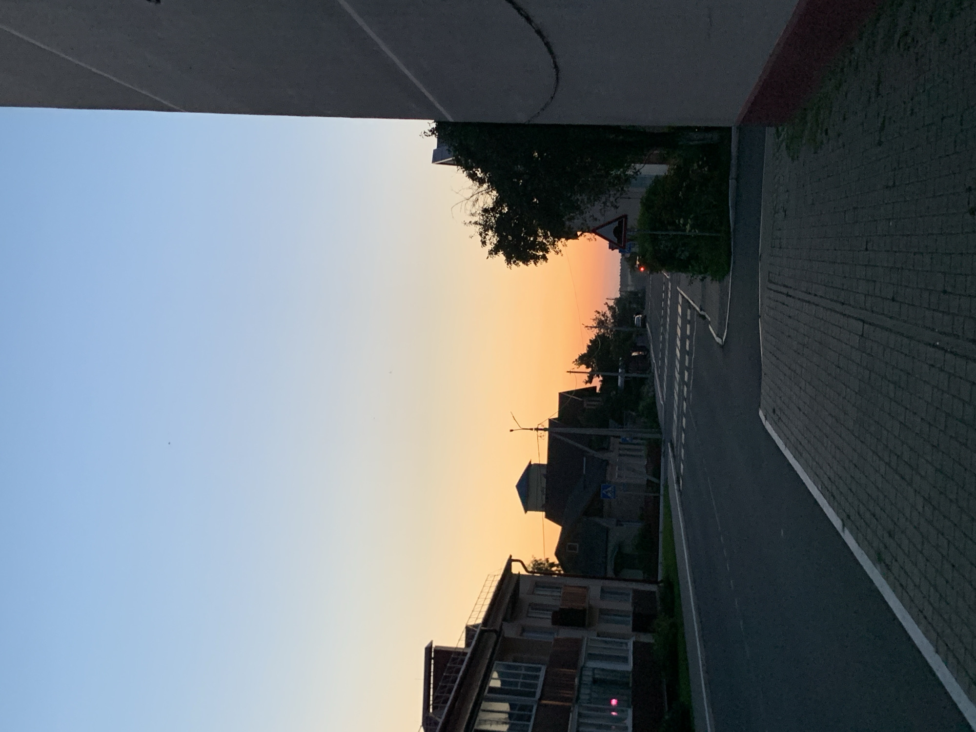 The image depicts a street scene in the city of Kapyĺ, Belarus, during twilight. On the left side of the street, a multi-story apartment building with several windows is visible. Across the street, there are houses with dark roofs and a pedestrian crossing. A traffic sign indicating a speed bump is present on the right side of the street, next to a densely vegetated area. The sky transitions from a pale blue at the top to an orange hue near the horizon. The street is paved, and a sidewalk made of grey bricks is in the foreground on the right. The right edge of the image is dominated by the side of a large building with a textured grey facade and a red base. The overall impression is a quiet, residential street at dusk.