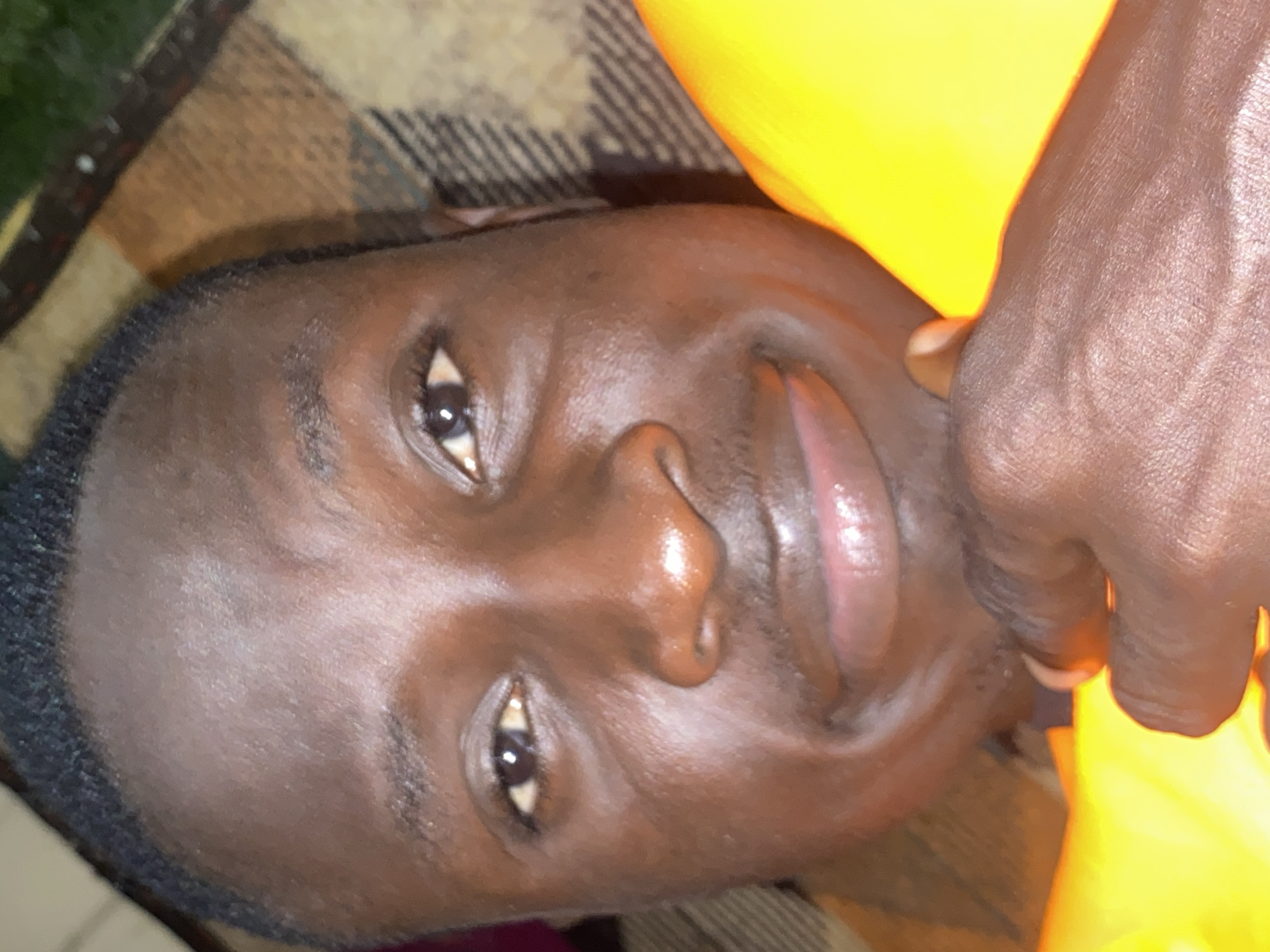 A close-up, head-and-shoulders shot captures a Black individual with dark skin and short, dark hair. The person is looking directly at the camera with a subtle smile, resting their chin on their left hand. A bright yellow object, possibly clothing or a life vest, occupies the right side of the frame. The background consists of a brown and beige patterned blanket. The image appears to be taken outdoors, with hints of green foliage visible in the upper right corner. The lighting suggests it could be daytime. The scene is set in Barden Yana, Nigeria.