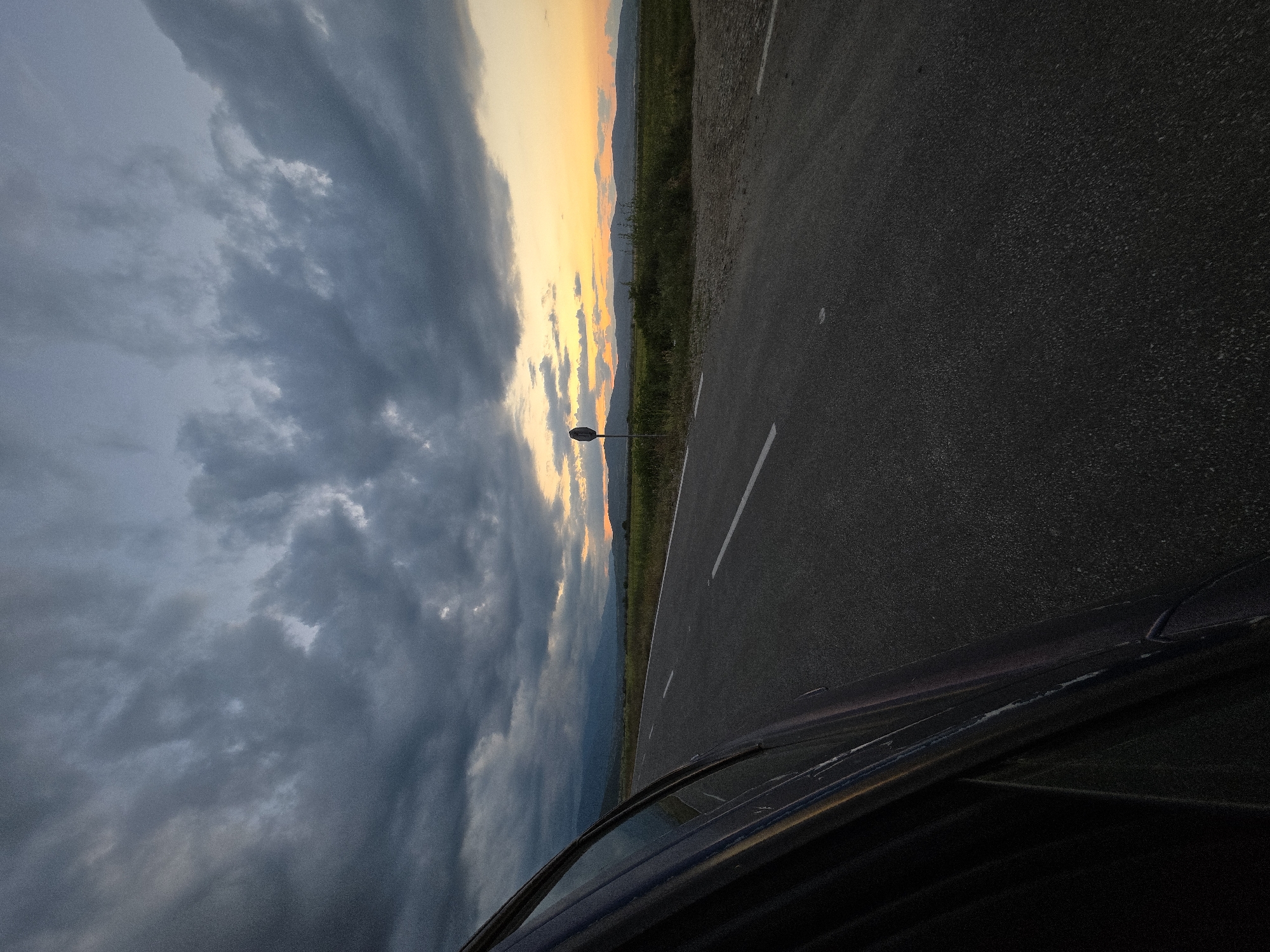 The image captures a rural road scene at dusk in Semchinovo, Bulgaria. The upper two-thirds of the frame are filled with heavy, dark gray cloud formations, which yield to a vibrant orange and yellow sunset glow on the horizon to the right. A dark asphalt road, marked with white dashed center lines and a solid white line on the right shoulder, extends into the distance, appearing to curve slightly left. The left side of a dark-colored vehicle, from which the photograph was taken, occupies the left foreground. Adjacent to the road on the right, a circular road sign on a pole is visible, positioned before an expanse of green fields or scrubland. A range of mountains is silhouetted against the lighter horizon in the background. No individuals are present in the frame.