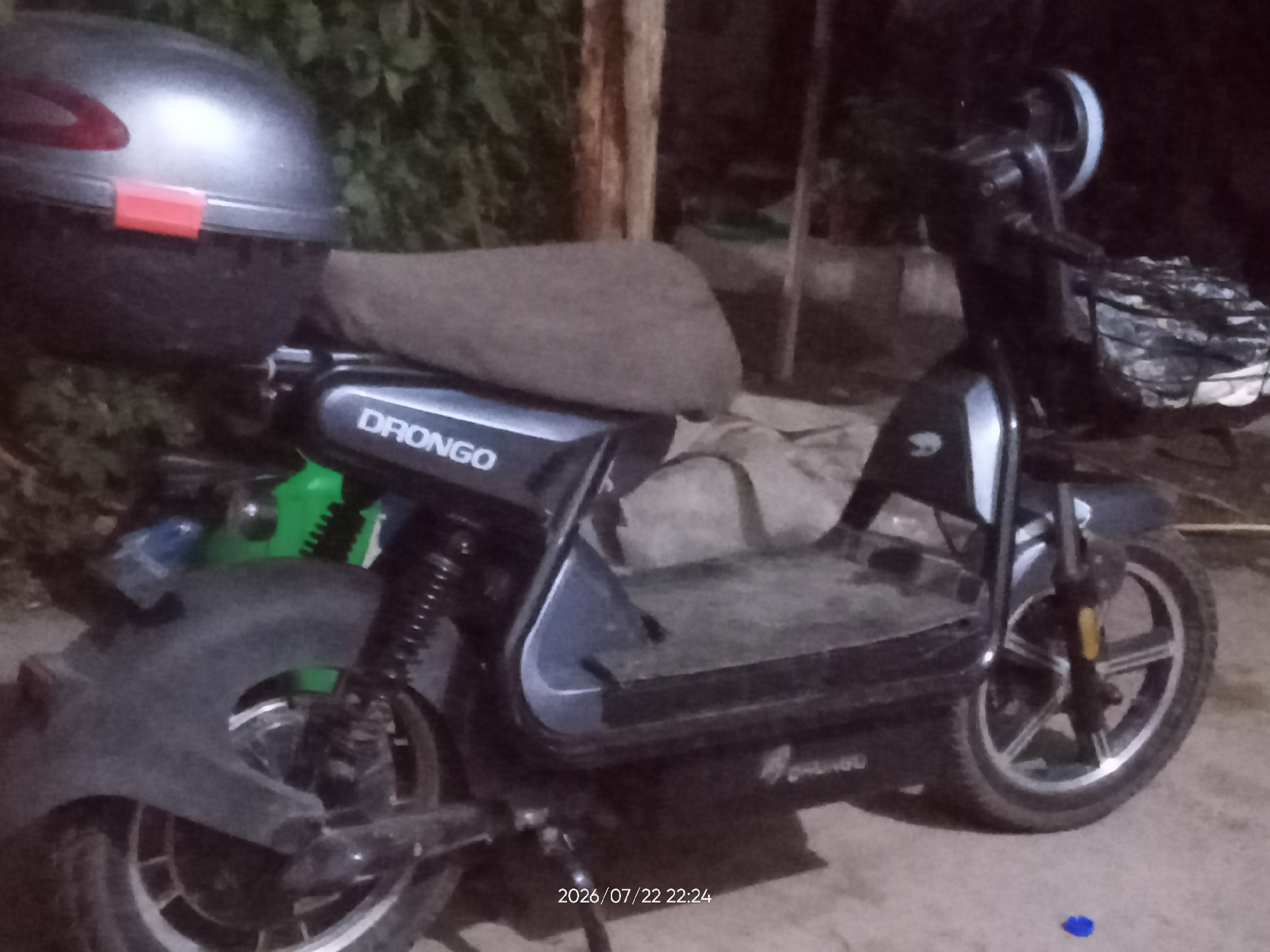 A parked black electric scooter, branded "DRONGO", is visible in an outdoor setting. The scooter is equipped with a large storage box on the rear and a basket on the front. The scene appears to be nighttime or low-light conditions, as indicated by the timestamp "2026/07/22 22:24" and the general darkness of the environment. There are hints of greenery and a wooden post in the background, suggesting an urban or suburban area. No people are visible in the image. The scooter is positioned on a paved surface.