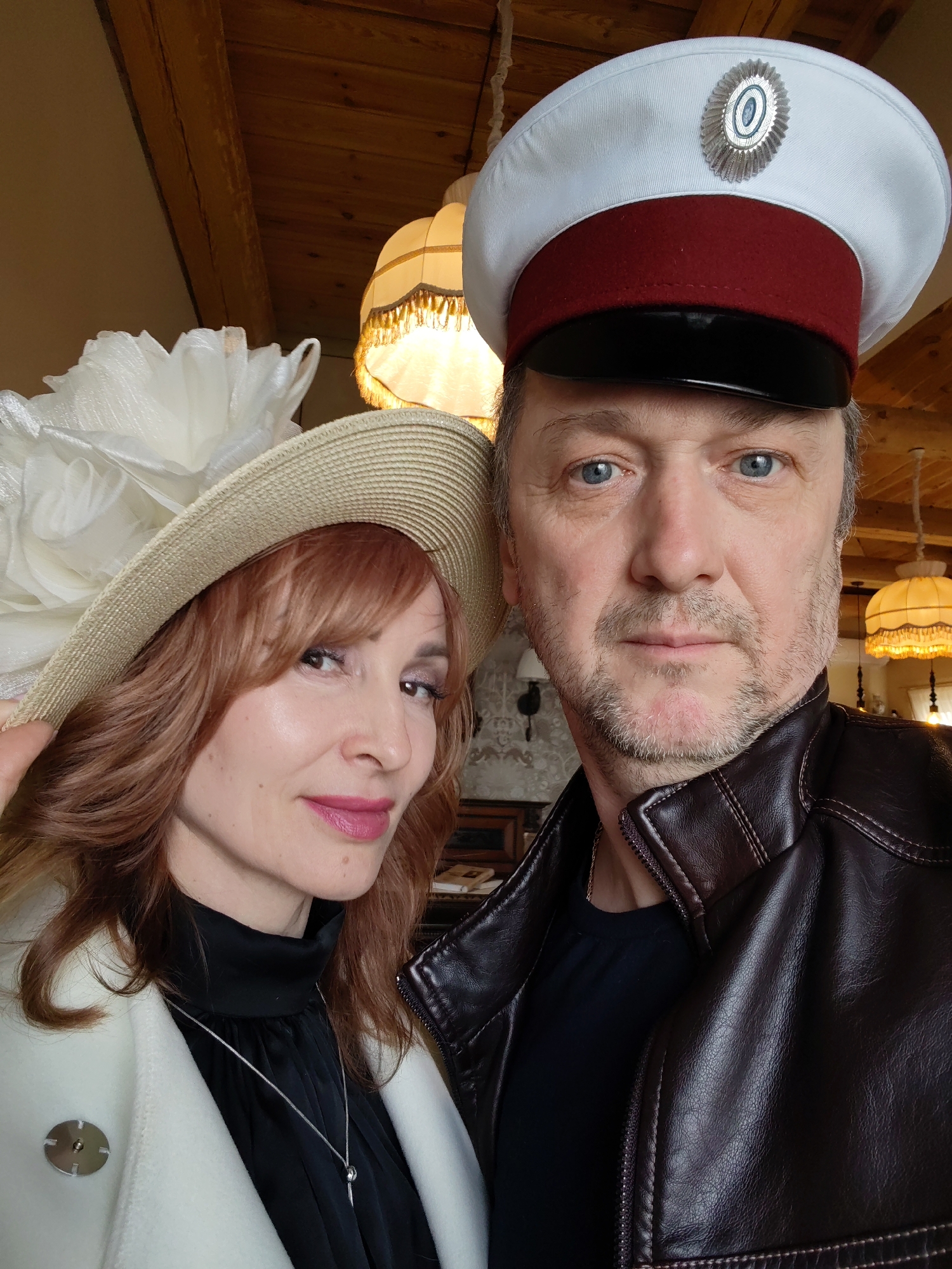 The photograph shows a man and a woman posing together. The woman is wearing a light-colored hat with a large white floral embellishment, a white coat, and a black top with a necklace. She has auburn hair and is wearing lipstick. The man is wearing a white and red cap with a badge, and a brown leather jacket over a dark shirt. The background appears to be an interior setting with lamps and wooden ceiling beams visible. There are also hints of furniture in the background. The location of the photograph in Sarapul, Russia, is not definitively indicated by any elements present in the image.