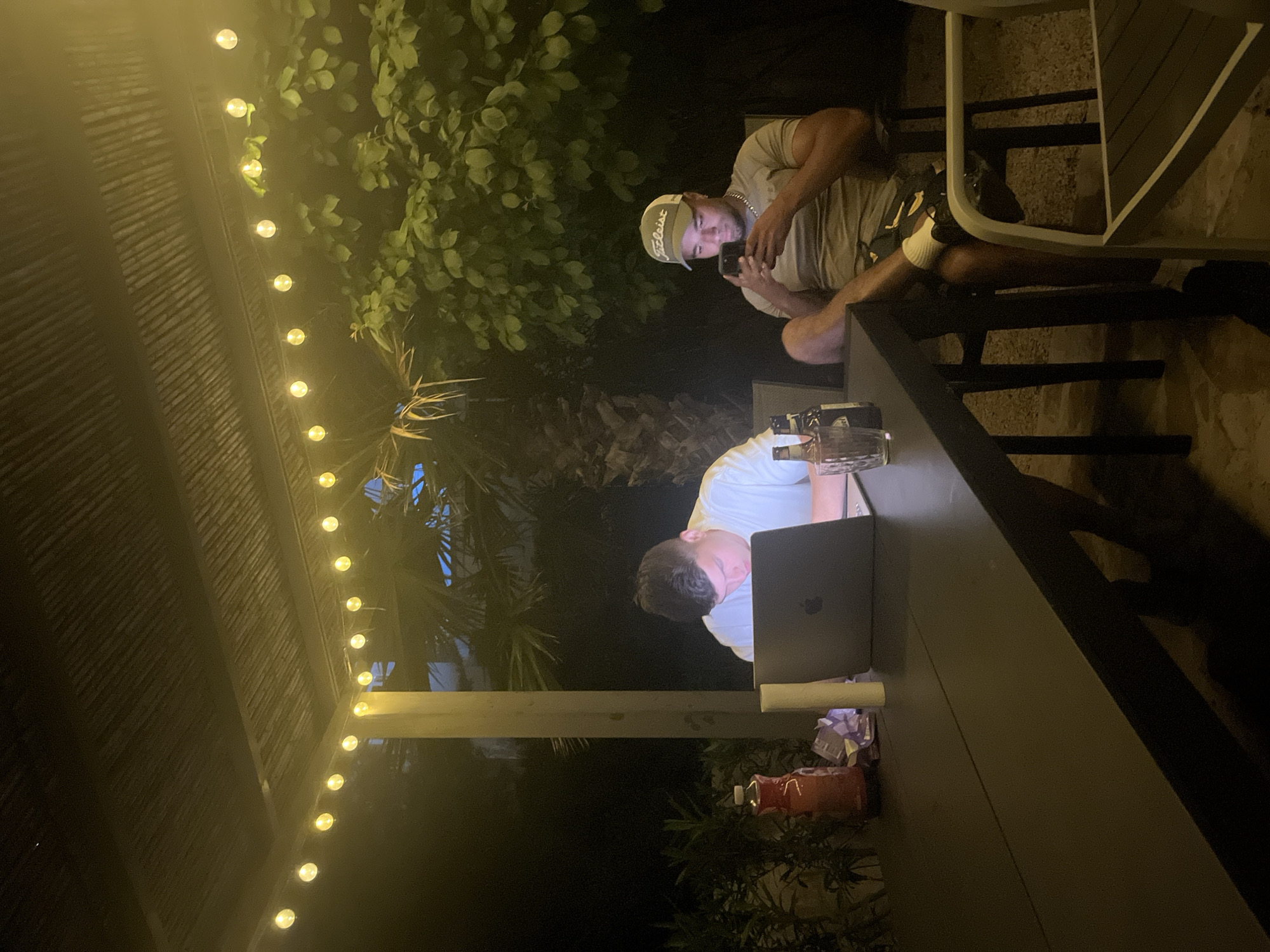 The image captures an outdoor scene at night, likely in a backyard or patio setting in el Poble Nou de Benitatxell / Benitachell, Spain. Two individuals are present: one is seated in a chair, looking at their phone, wearing a light-colored cap and a grey t-shirt. The second person is seated at a long, dark table, working on a laptop. Various objects are on the table, including a glass bottle and other containers. String lights are visible overhead, illuminating the area, and lush greenery surrounds the patio.