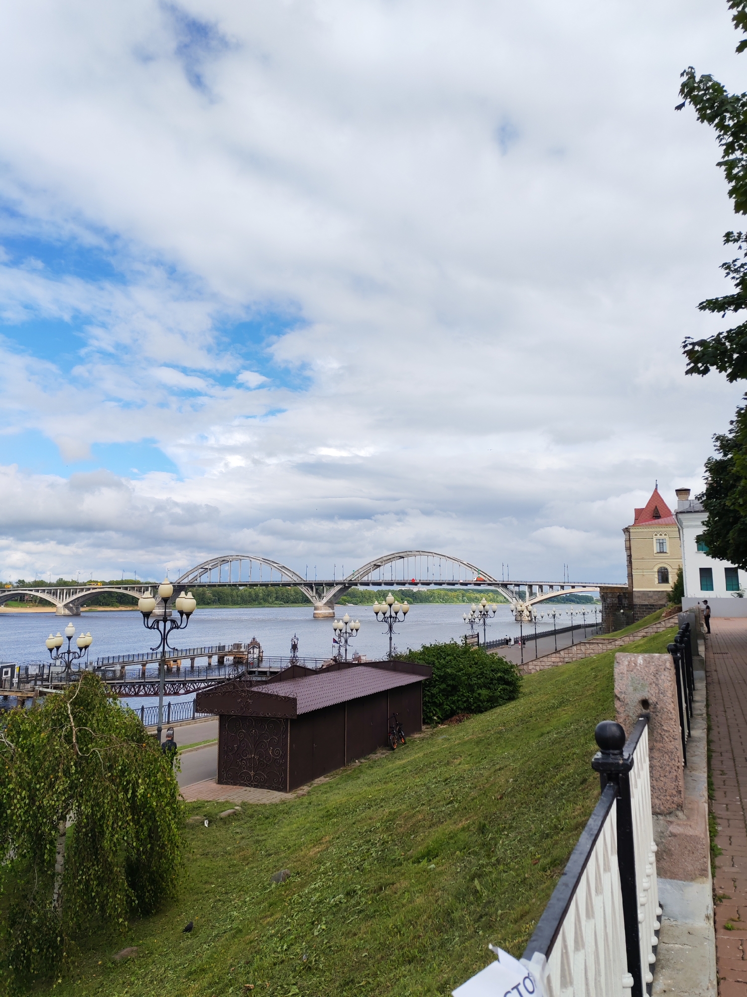 The image shows a wide, calm river with two bridges spanning across it. The bridge on the left is a long, multi-arched structure, while the bridge on the right features a prominent, high arch. The riverbank on the right side of the image is lined with a paved walkway and a low black metal fence. Beyond the walkway, there is a grassy embankment dotted with trees and a few buildings, including a distinctive building with a red-tiled roof. Several streetlights are visible along the walkway. The sky is partly cloudy, with patches of blue sky visible through the white clouds. A lone person is standing on the walkway near the building with the red roof, facing away from the camera and towards the river. The overall scene depicts a tranquil riverside urban landscape. This location appears to be the embankment of the Volga River in the city of Rybinsk, Yaroslavl Oblast, Russia, featuring the Rybinsk Automobile Bridge and the Old Volga Bridge.