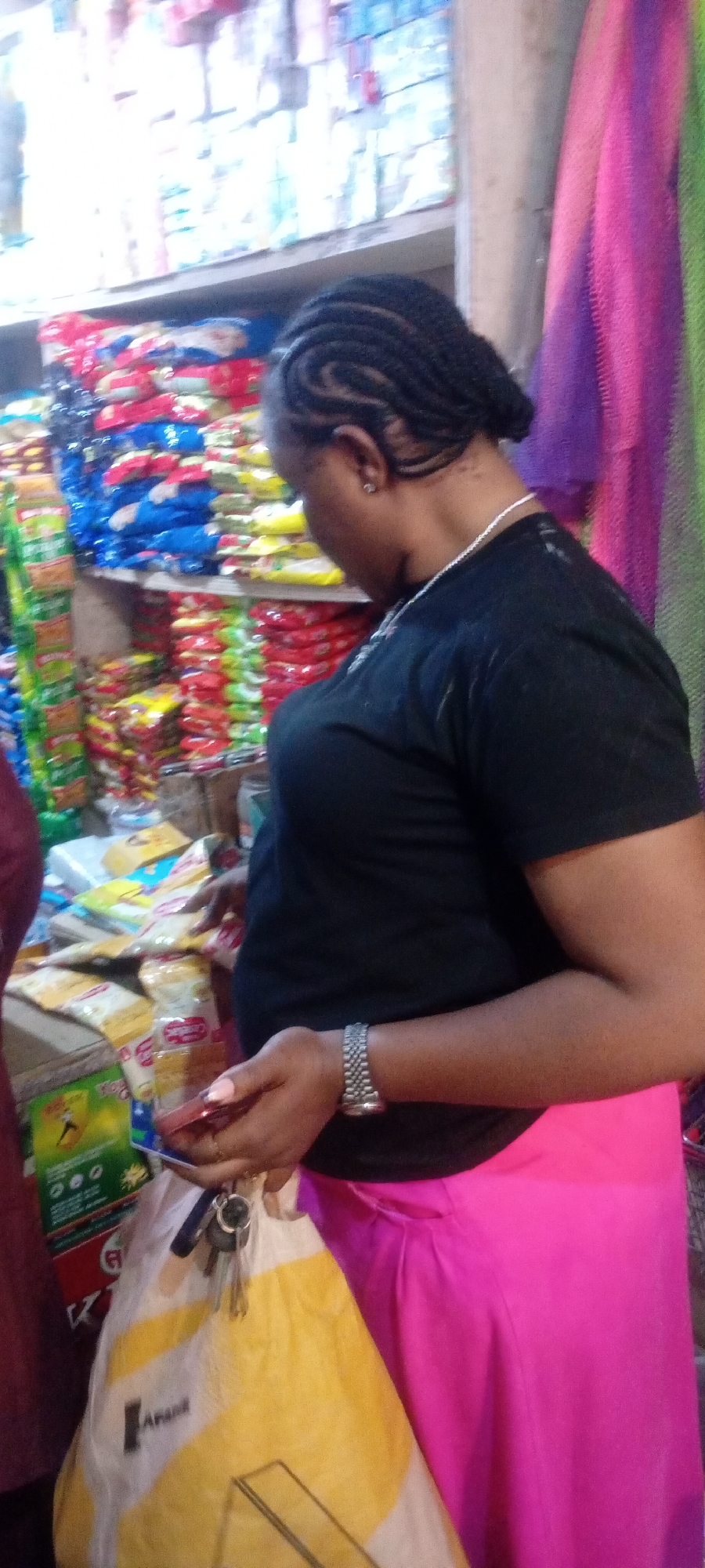 A woman is shopping in a store in Kano, Nigeria. She is wearing a black T-shirt and a bright pink skirt. Her hair is braided and pulled back. She is holding a package of something in her hand, examining it. A yellow and white shopping bag hangs off her wrist with keys attached. The shelves behind her are filled with various food items and household goods. Multicolored fabric hangs on the right side of the frame.