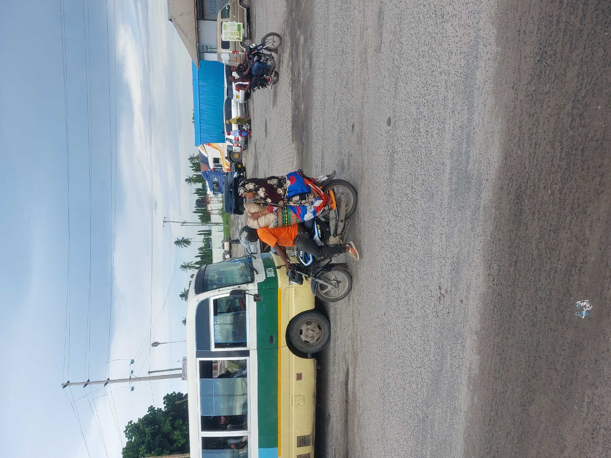 The image depicts a scene on a paved road in Dar es-Salaam, Tanzania. A yellow and green bus is parked on the left side of the road. A motorcyclist wearing an orange shirt and helmet is driving next to the bus with a female passenger on the back. Further back, another motorcycle with two people is parked near several other vehicles. The weather is fair with a partly cloudy sky. Overhead power lines are visible on the left of the image.