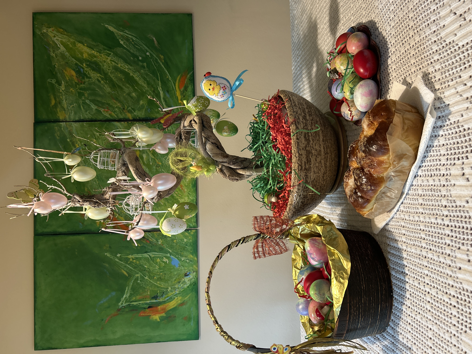 A still life image shows a table or sideboard with various Easter decorations against a wall in an indoor setting. A large green abstract painting composed of three panels hangs on the wall behind the arrangement. The painting's style suggests the scene could be taking place in a home in Unknown, Unknown.
On the table, from left to right:
1. An Easter egg tree stands tall, adorned with pastel-colored eggs, small cages, and butterfly decorations. It appears to be a decorative tree, possibly made of branches, displayed as an Easter centerpiece.
2. A textured pot filled with green and red shredded paper simulating grass holds a decorative Easter chick on a stick.
3. A shallow dish containing a collection of colorful Easter eggs, with a variety of colors and patterns.
4. A loaf of bread, possibly a sweet bread traditionally served during Easter, is partially wrapped in white parchment paper.
5. A brown basket with a golden lining, filled with Easter eggs, is topped with a ribbon bow and has a small yellow decorative piece at its base.
The entire composition is set against a textured, light-colored tablecloth. The arrangement suggests a celebration of Easter, complete with decorative eggs, a themed centerpiece, and a festive bread.