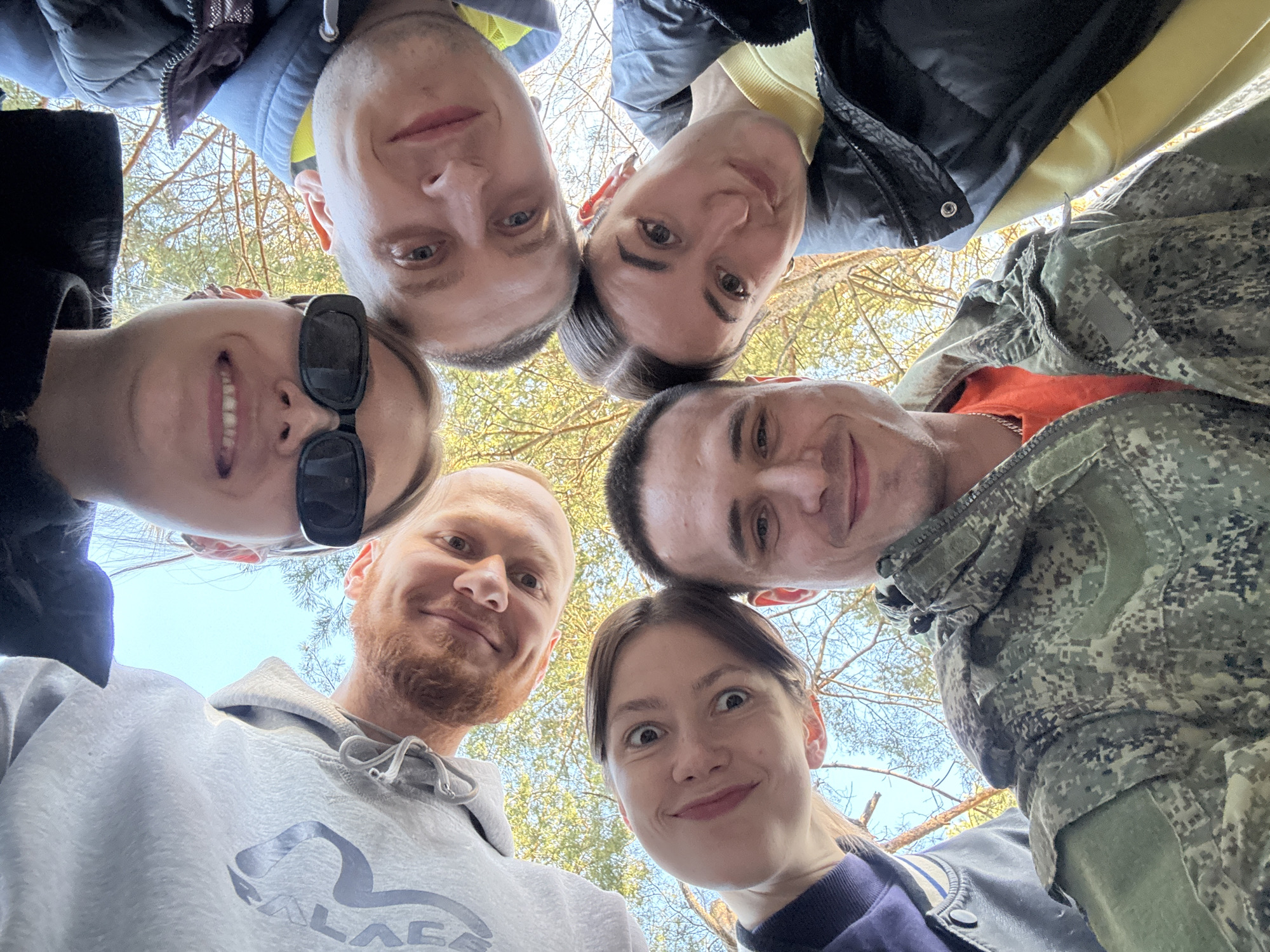 A low-angle shot captures six people looking down at the camera. The background reveals a pale blue sky with the silhouettes of tree branches. The people's clothing varies, including a grey sweatshirt, a camouflage jacket, a black and yellow jacket, and a dark-colored jacket. One person wears sunglasses. The individuals appear to be in a forest or park. This is a picture of a group of friends taken in Chaikovsky, Russia.