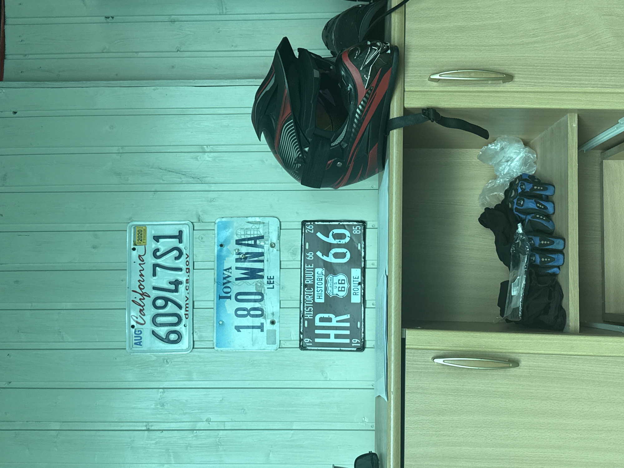 The image depicts a wall with vertical wood paneling, painted in a teal hue. Three license plates are mounted on the wall. The top plate is from California, with the number "60947S1" and "dmv.ca.gov" printed on it. Below that is an Iowa license plate reading "180 WNA LEE". The bottom plate is from Oklahoma, a "HISTORIC ROUTE 66" plate with "HR 66" and "ROUTE" printed on it. To the right of the license plates, a black and red motocross helmet is placed on a light-colored cabinet. On the shelf of the cabinet, several pairs of blue and black gloves and some plastic-wrapped items are visible. The overall setting appears to be an indoor storage area or garage. The image does not provide any information about the city of Novokuybyshevsk, Russia.