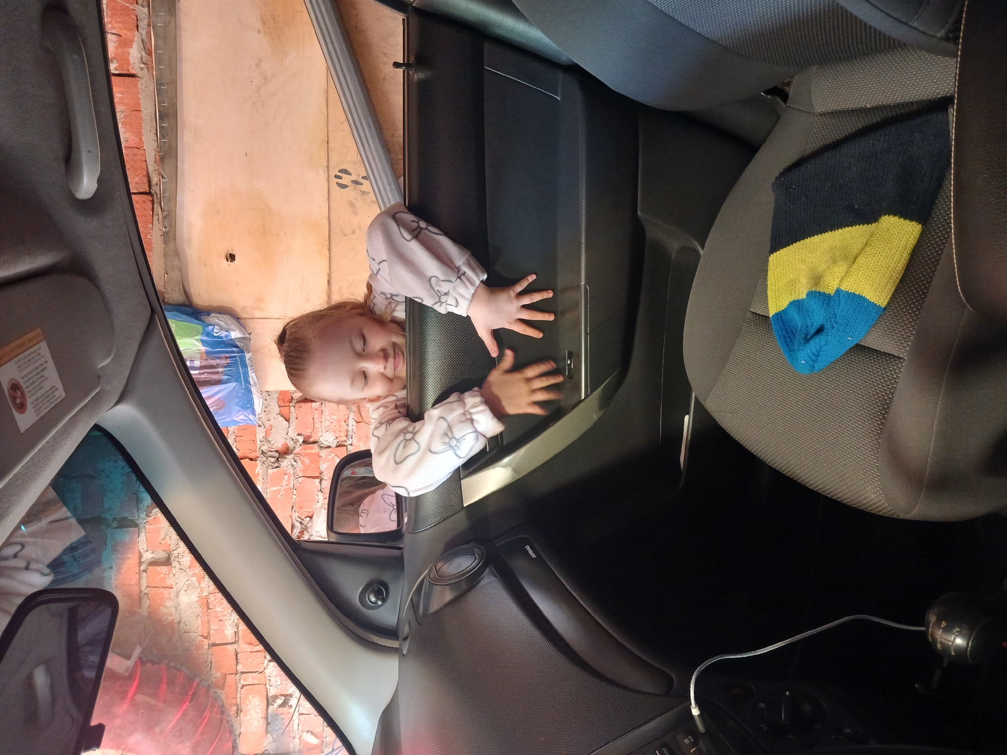 A young girl is lying in the opening of a car door, with her hands resting on the door panel. She is wearing a light-colored pajama top with a bow pattern. The car's interior is visible, including the dashboard, seats, and a sock on the passenger seat. The exterior shows a brick wall and a garage door. There is no evidence to confirm the location is in Noviy, Russia.