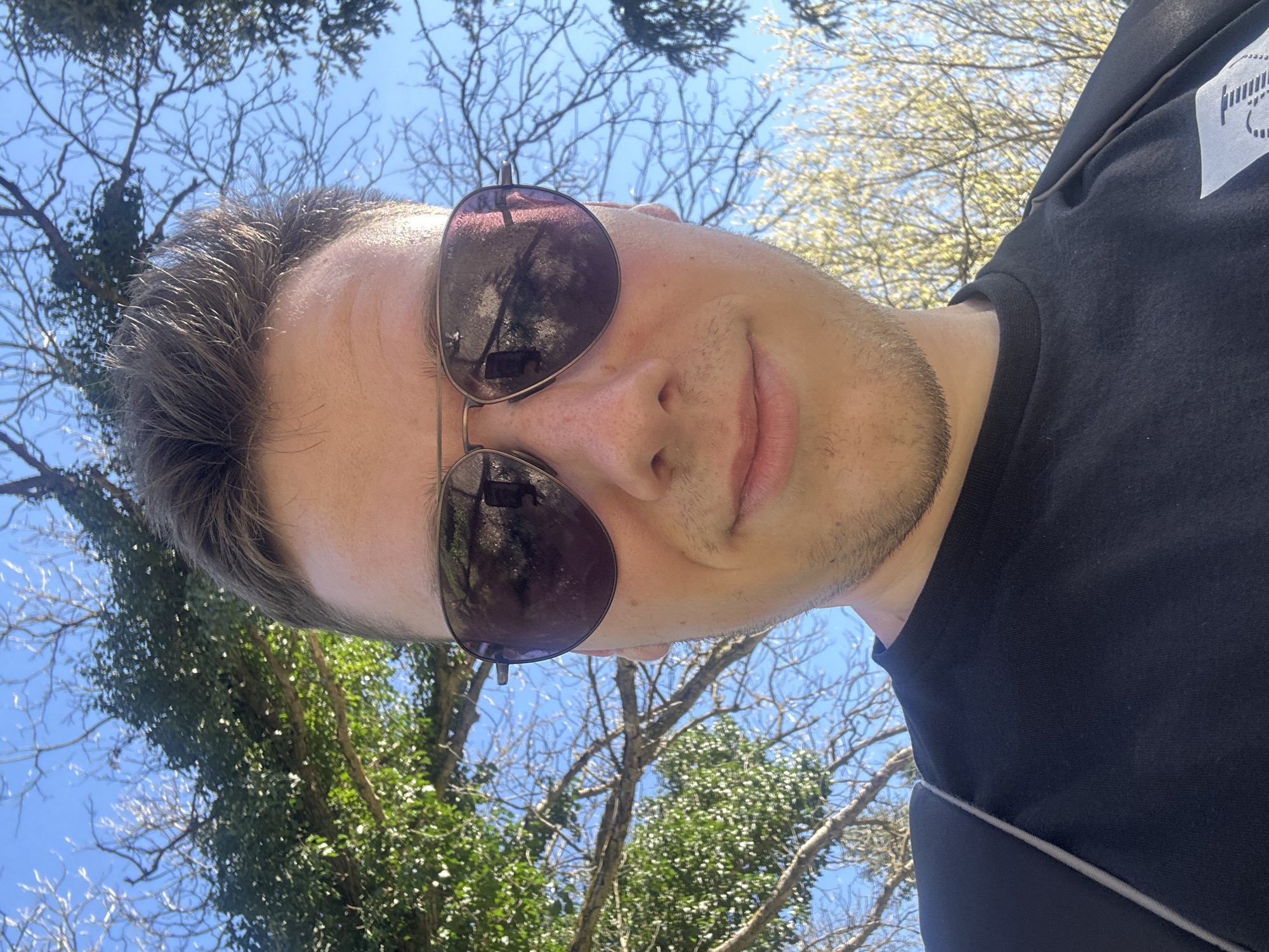 A selfie of a fair-skinned person with short, dark hair, wearing aviator sunglasses and a black t-shirt, is taken outdoors under a blue sky with trees visible in the background. The person has stubble and is looking slightly off-center. The sunglasses reflect the surrounding trees. The photo is taken at an upward angle. The location is Pleven, Bulgaria.