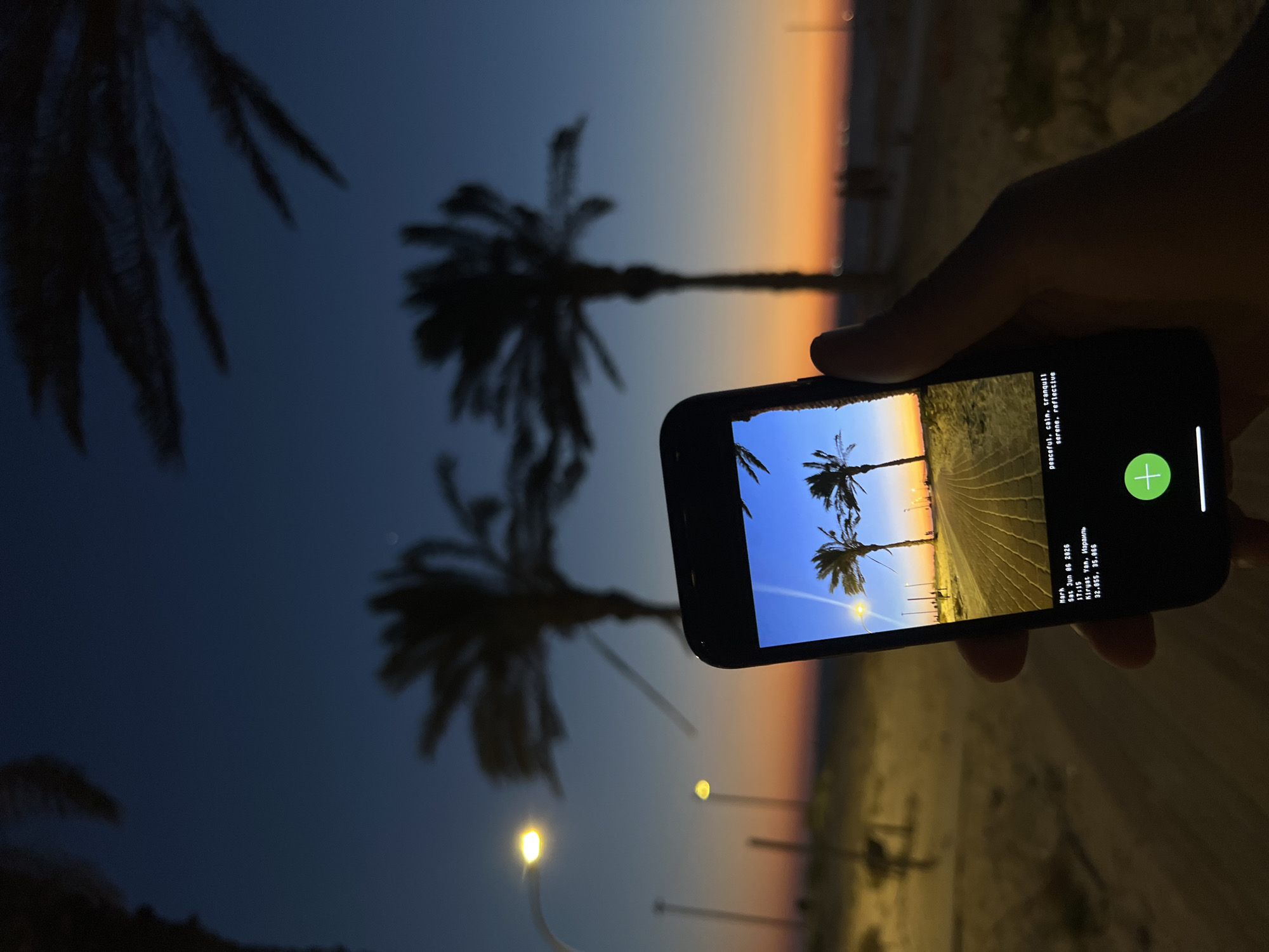 A right hand holds a dark smartphone, presenting a "photo within a photo" composition. The smartphone screen displays an image of a paved path leading towards the horizon, flanked by two silhouetted palm trees against a sunset sky transitioning from blue to orange and yellow. A street lamp illuminates the left side of the path within the screen's image. Text overlay on the screen identifies the displayed photo as taken on "Sat Jun 08 2026" at "17:15" in "Kiryat Yam, Israel" (coordinates 32.899, 35.066), accompanied by descriptive tags such as "peaceful, calm, tranquil."
The actual background scene mirrors the content on the phone screen, depicting a sunset over a coastal area in Kiryat Yam, Israel. Silhouetted palm trees frame the upper and right portions of the sky, which exhibits a deep blue overhead gradually blending into warm orange and yellow hues closer to the horizon. Two streetlights are visible, emitting bright yellow light on the left side of the scene. The foreground and midground consist of a dark, sandy beach or promenade area. Faint, blurry figures and distant lights are discernible along the horizon line.