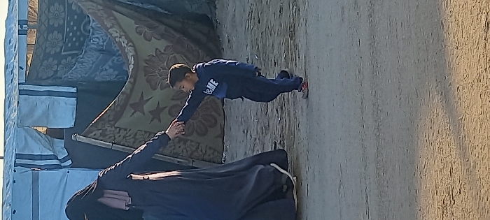 In Khan Yunis, Palestinian Territory, a woman in a dark abaya and black head covering is depicted holding the left hand of a young boy. The woman is partially visible on the left, with a light pink under-scarf around her neck. The boy, positioned centrally and to the right, wears a dark blue tracksuit with "B&ME" visible in white text on his hoodie, and light-colored shoes with orange accents. He extends his arm, holding the woman's hand, and appears to be stepping forward. The subjects are situated on an uneven, light brown dirt or sand ground. In the background, makeshift shelters are constructed from various materials, including a light blue or white tarp with vertical blue stripes on the left and center, and patterned blankets or rugs, one displaying floral motifs and two stars, on the right. A clear, light blue sky is visible at the top. Natural light illuminates the scene.
