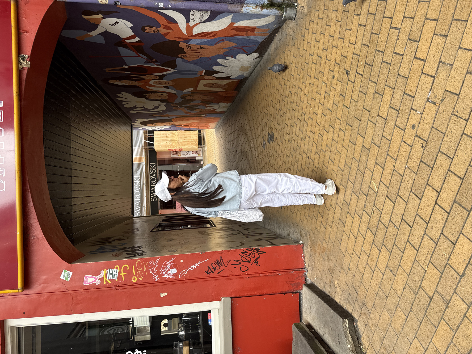 A woman wearing a white cap, a light blue denim jacket, and white pants is walking away from the camera down a narrow alleyway with a yellow brick floor. She is carrying a white tote bag over her left shoulder and appears to be holding a cigarette in her right hand near her mouth. The alleyway has a red painted wall on the left, adorned with graffiti, and a vibrant mural on the right wall depicting a crowd of people. Above the mural, the word "SWAROVSKI" is visible on a sign. A pigeon is seen on the ground in the middle of the alleyway. The scene appears to be in the city of Groningen, Netherlands, specifically within an alleyway that leads to the Grote Markt.