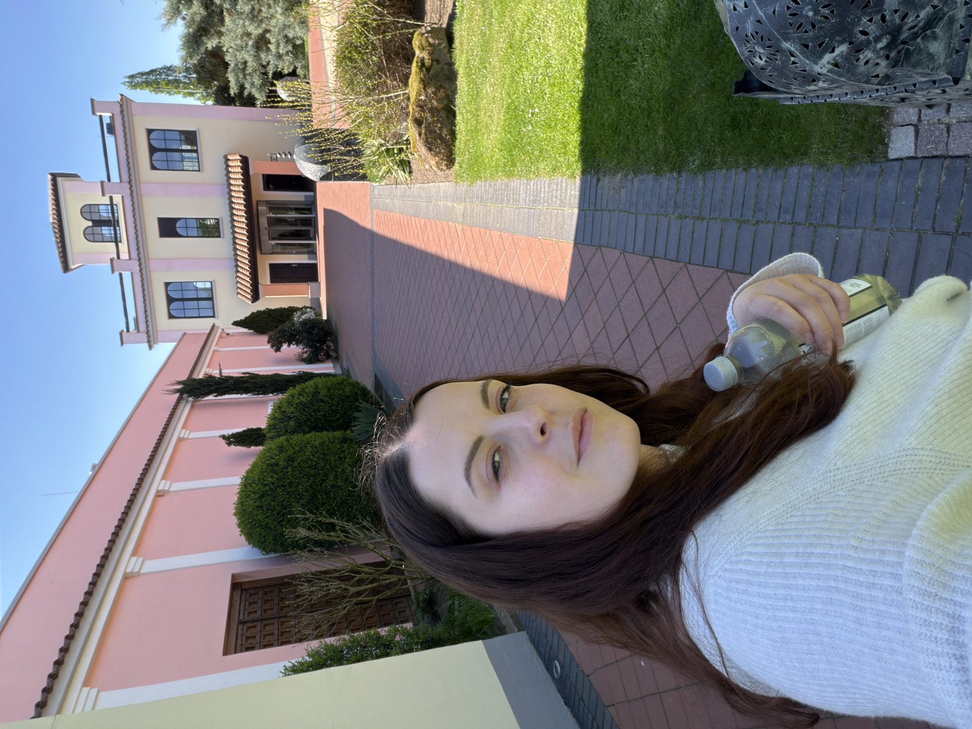 This outdoor selfie, set in Bergisch Gladbach, Germany, captures a young woman in the foreground with a distinct architectural building and manicured landscaping behind her. The photograph is taken on a bright, sunny day, indicated by the clear blue sky and strong shadows cast on the paved ground.
The primary subject is a woman with long, dark brown hair, looking towards the camera with a calm, neutral expression. She is wearing a light-colored, possibly white or light grey, knit sweater. Her right hand is visible, holding a clear plastic water bottle, though any text on its label is indistinct.
In the mid-ground, a multi-story building with light beige walls and decorative pink trim around its windows and eaves is prominent. The building features dark-framed windows, some of which have arched tops, adding to its traditional European aesthetic. A main entrance with dark double doors is visible beneath an awning. To the left, a section of another building with similar pink-toned walls can be seen.
The outdoor setting includes a vibrant green lawn to the right of the paved area, flanked by neatly trimmed bushes and some sparse tree branches. A large, dark, ornate metal garden lantern or sculpture is partially visible on the far right. The ground is covered with a patterned red-brick path that transitions into sections of darker grey/blue paving stones, creating a well-maintained courtyard or garden pathway. The overall scene suggests a tranquil, well-kept property, possibly a hotel or guesthouse.