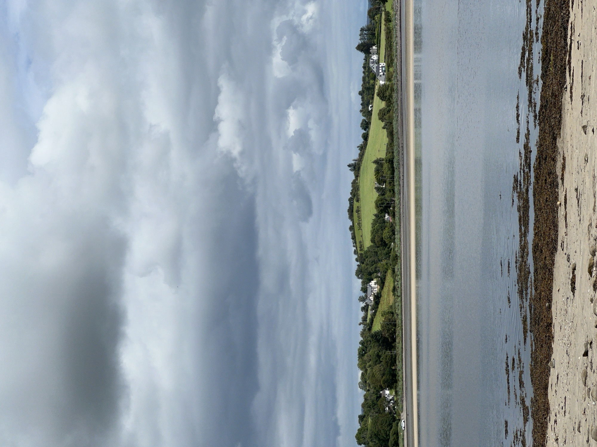 The image shows a coastal landscape in Mountcharles, Ireland, under a heavily clouded sky. In the foreground, a sandy beach is visible, scattered with small rocks and dark seaweed. A body of water, likely a bay or estuary, occupies the midground. The water's surface is calm, with ripples reflecting the overcast sky. Across the water, a lush green hillside rises, dotted with several white houses and trees. A road runs along the base of the hill, with a white car visible on it. The overall impression is one of a tranquil, rural scene on a cloudy day.
