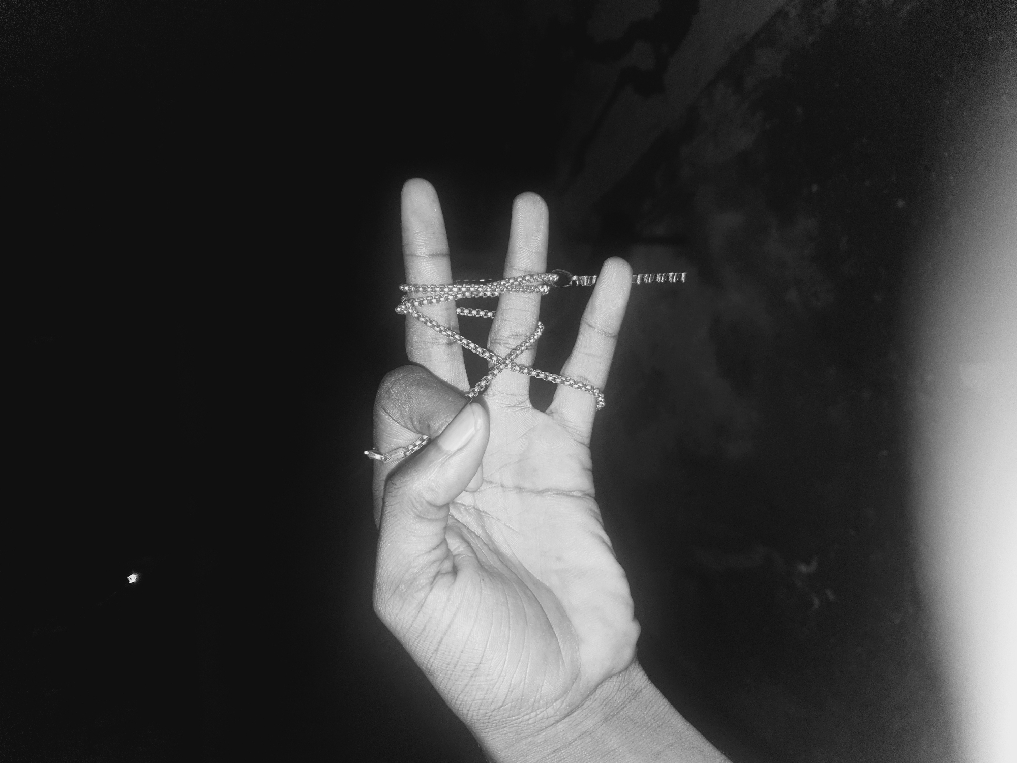 A black and white photograph shows a hand with a silver chain wrapped around its fingers. The hand is positioned in the center of the frame, and the background is dark. The chain appears to be a thin, linked metal chain. The setting is not clear enough to identify the location in Lokoja, Nigeria.