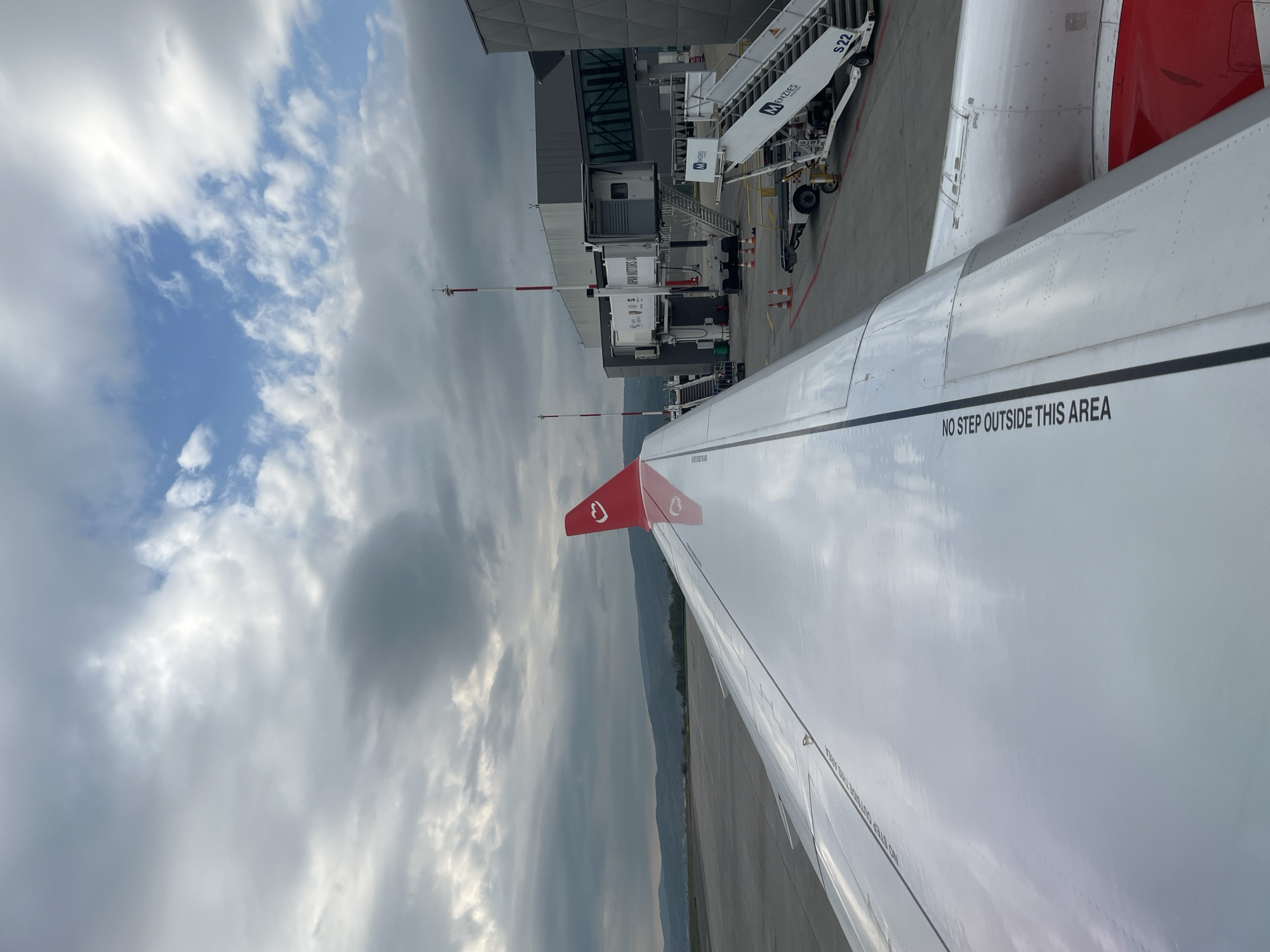 A view from an airplane window shows a portion of the wing with the warning "NO STEP OUTSIDE THIS AREA". The tail fin, painted red with a white emblem, is also visible. Beyond the wing, there's a jet bridge and a set of stairs with "MENZIES" branding. The scene suggests the airplane is parked at an airport terminal. Beyond the terminal infrastructure is a landscape with hills under a sky with a mix of blue and grey clouds. Location: Unknown, Unknown.