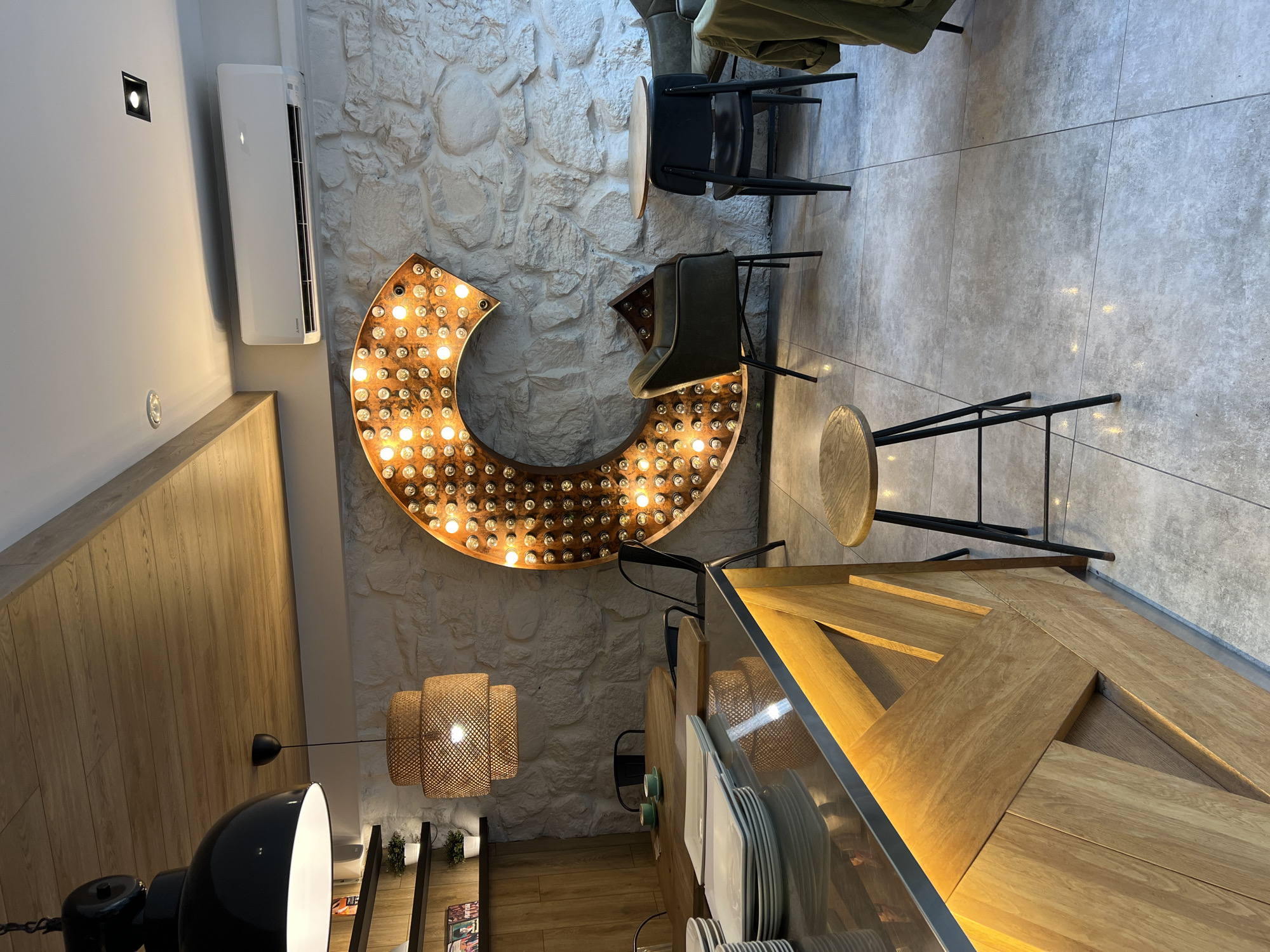 The image depicts the interior of a modern cafe, potentially located in Palanga, Lithuania. In the foreground, a polished dark countertop of a bar or service area is visible, illuminated from below by warm-toned LED lighting. The front of the bar features a distinctive wooden design with angled planks. To the left of the bar, a stack of white square plates is neatly arranged.
In the mid-ground, a large, stylized letter "C" made of illuminated bulbs is prominently displayed against a textured stone wall. To the right of the "C," a small round table with two black chairs sits. Further right, another dark armchair and a glimpse of more seating suggest a seating area for patrons. A light-colored air conditioning unit is mounted high on the wall above the "C."
In the background, a round wooden table with two chairs and two green teacups is visible. Shelving units are mounted on a wood-paneled wall on the left, displaying books and small potted plants. Above the service area, a contemporary black pendant lamp hangs. A woven pendant lamp with a wicker shade is also present. The floor is tiled with large, light gray tiles. The overall ambiance is contemporary and inviting, with a mix of natural materials and industrial-style lighting. No people are present in the scene.