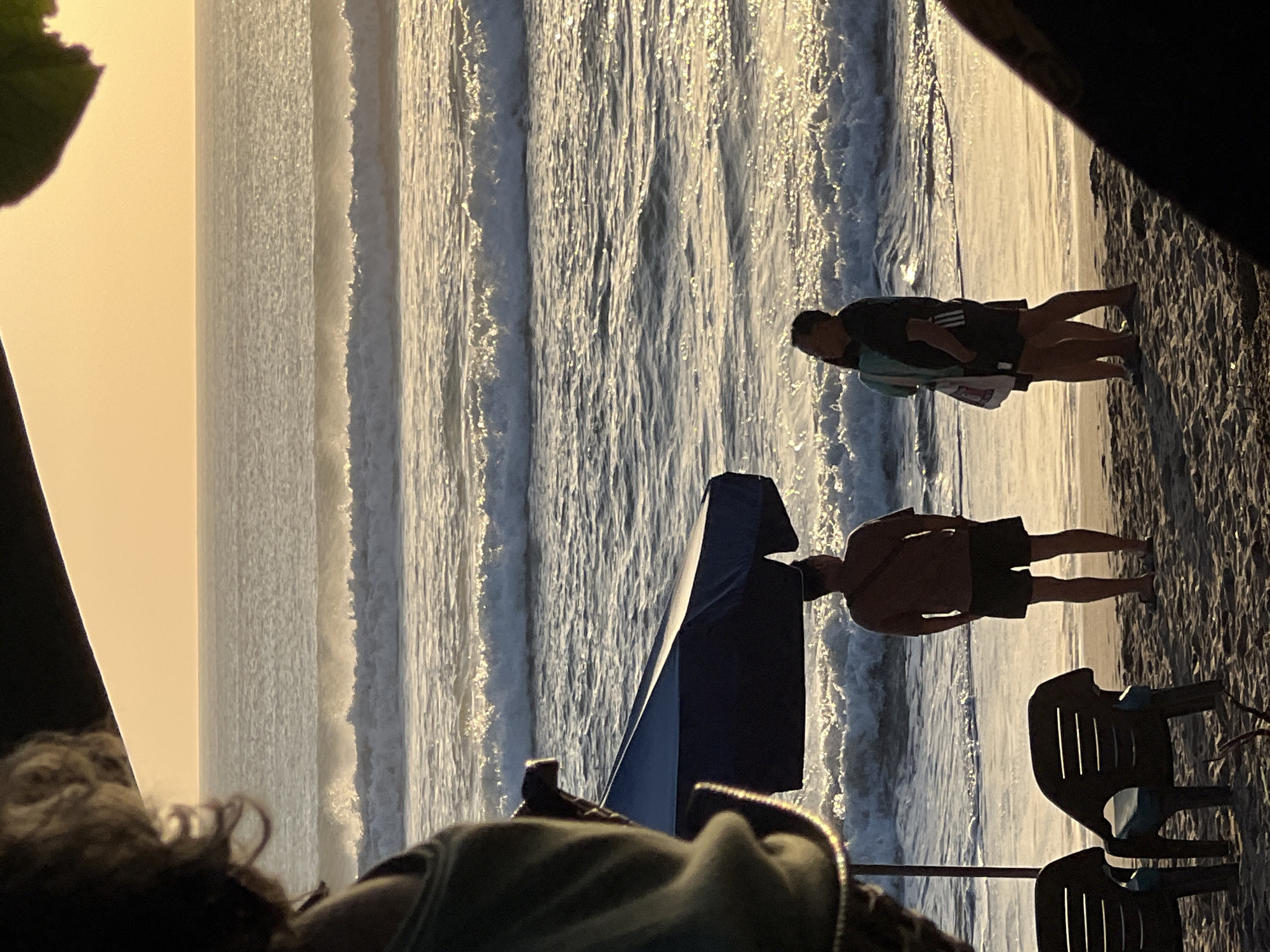 An outdoor scene at dusk on Kuta Beach, Indonesia, shows the ocean with gentle waves and a golden sky. In the foreground, two empty plastic chairs are on the sand. A blue beach umbrella stands behind the chairs, shading two individuals. A man in a brown t-shirt and dark shorts stands facing the ocean, his hands in his pockets. To his right, a couple stands together; the woman in a light teal top and a man in a dark t-shirt and dark shorts. The man has his arm around the woman. Both individuals are also facing the ocean. The overall mood is relaxed and peaceful, with the warm light of the setting sun reflecting on the water.