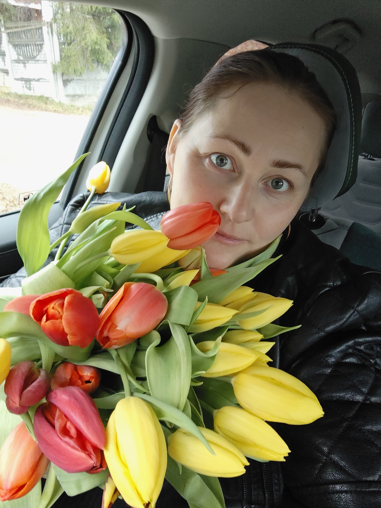 A woman is captured in a close-up portrait, possibly a selfie, seated inside a car in Markovo, Russia. She appears to be in her 30s or 40s, with fair skin, brown hair pulled back, and striking green-blue eyes that convey a direct, perhaps slightly surprised, gaze towards the camera. She is wearing a black quilted jacket.
A large, vibrant bouquet of fresh tulips dominates the foreground, held by the woman. The tulips are a mix of red, orange, and bright yellow, with prominent green leaves and stems. Some of the tulip buds gently touch her chin and lips, suggesting the flowers are held close to her face.
The setting is the interior of a car, with a dark headrest visible behind her and a window frame to her left. Through the blurred car window, an outdoor scene indicates daytime under an overcast sky. Elements like a grey fence or wall and bare trees are faintly discernible, hinting at a suburban or semi-rural backdrop. The soft, diffuse lighting is consistent with cloudy weather. No visible text is present in the image. The scene suggests the woman has either just received or is transporting the fresh bouquet of tulips.