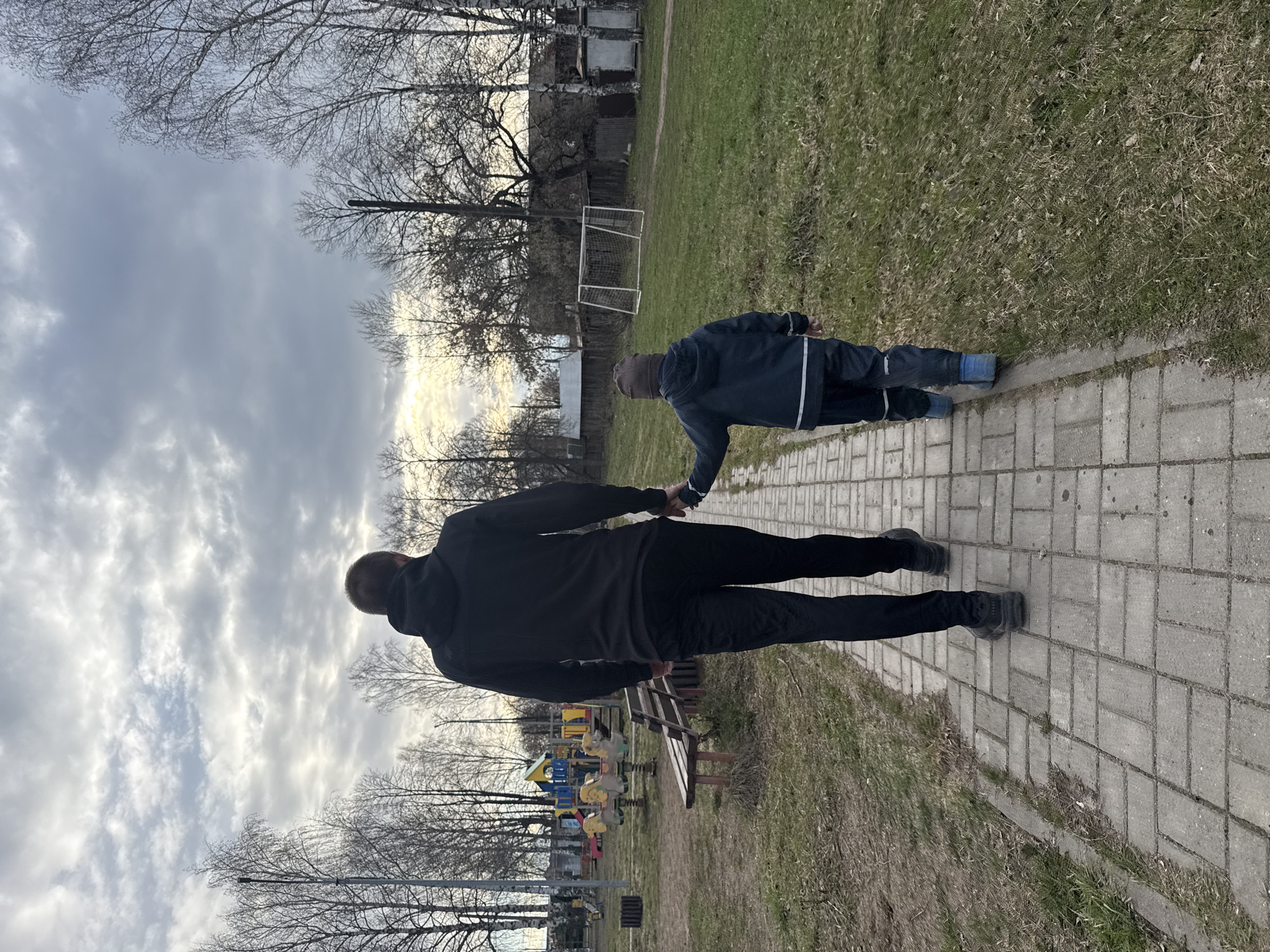 The image, correctly oriented, depicts an adult and a child interacting in an outdoor recreational area. In the foreground, a paved path made of gray rectangular blocks runs parallel to a grassy patch with sparse, dry grass.
An adult, viewed from behind, stands on the paved path. They are dressed in a black hooded jacket, black pants, and dark shoes. To their right, in the grassy area, a child is crouched on their hands and knees, facing away from the camera. The child wears a dark blue jacket with white stripes on the arms, dark pants, and blue boots, topped with a dark cap. The adult is holding the child's hand, suggesting a moment of support or encouragement.
In the midground, behind the adult, a colorful playground structure with slides and spring riders is visible, along with a wooden park bench. Further right, behind the child, a white metal soccer goal stands on the grass. The background is filled with numerous bare trees, indicating a late winter or early spring season. Some distant buildings are faintly visible behind the trees and goal. The sky is predominantly overcast, but brighter patches suggest the presence of the sun, possibly indicating late afternoon or early evening. The scene is consistent with a park or public outdoor space in Chaikovsky, Russia. No text is visible in the image.