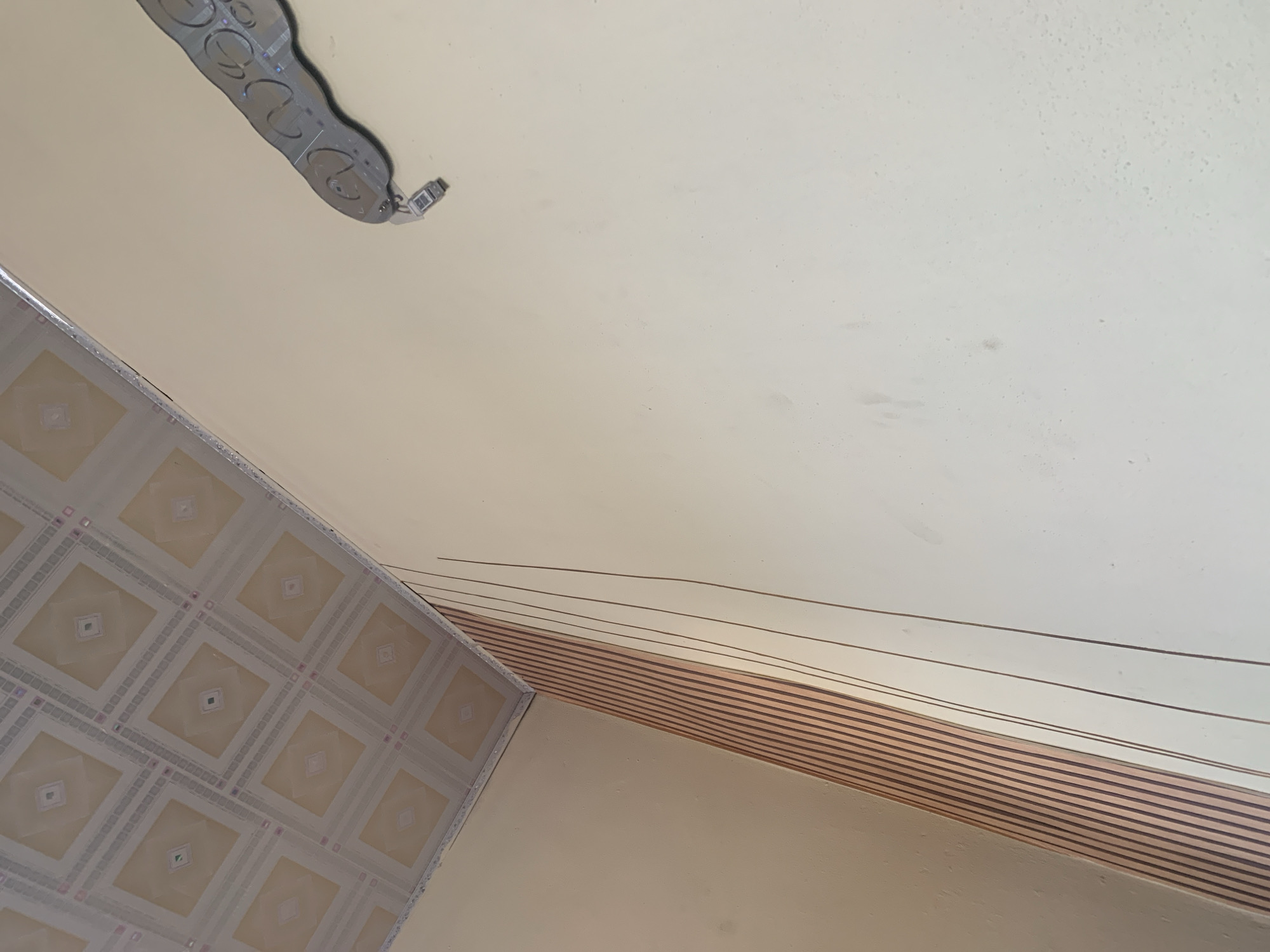 The image shows an interior view of a room. The ceiling is a tiled drop ceiling with a patterned design. On the right side of the image, mounted on the wall, is a silver object with multiple circular indentations. On the left side, the wall is adorned with vertical wooden slats, transitioning into a plain cream-colored wall. Several thin, dark lines run vertically down the cream wall from the ceiling. This scene is not located in Jauro Yinu, Nigeria, but appears to be a generic interior setting.