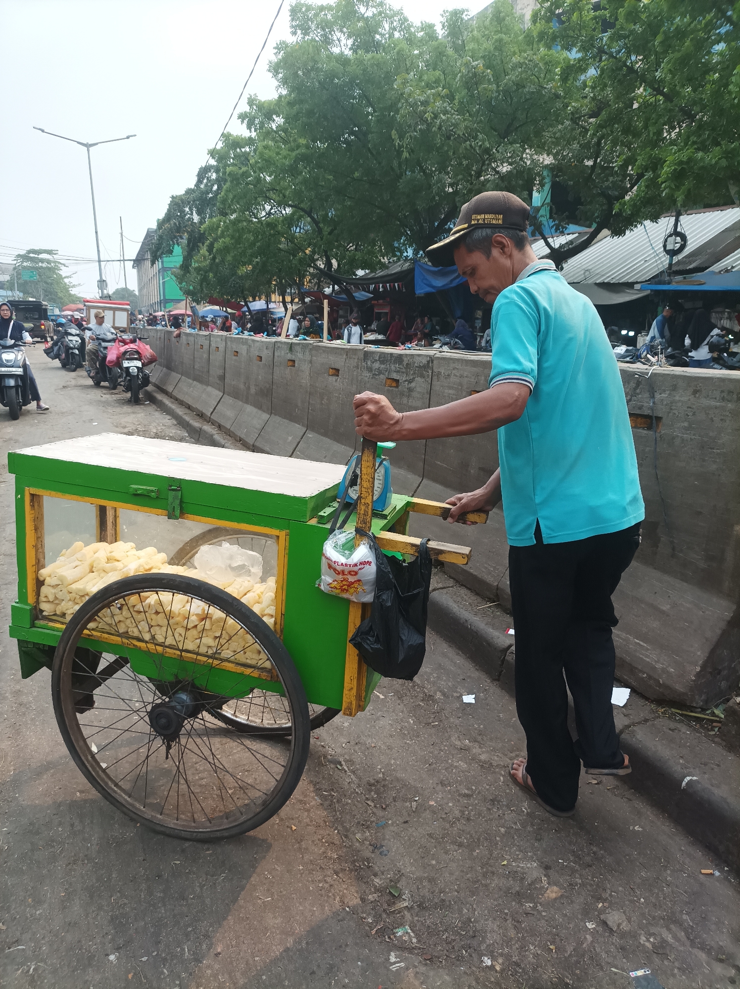 A street vendor in South Jakarta, Indonesia, is pushing a food cart filled with what appear to be fried cassava sticks. The vendor, a man wearing a blue polo shirt and dark pants, is seen from the side, with his gaze directed downwards. The food cart is green and yellow, with a glass display case showcasing the food. Behind the cart, a concrete barrier separates the street from a marketplace with various stalls and tents. Motorbikes are lined up on the street, indicating traffic or a parking area. The overall scene depicts a bustling urban environment with commercial activity.