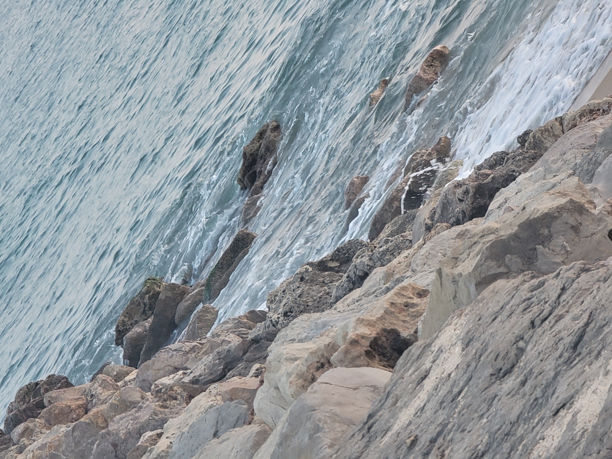 The image depicts a rocky coastline with the ocean meeting the shore. The foreground is dominated by large, grey and beige rocks that form a rugged barrier. These rocks are varied in size and texture, with some appearing weathered and others more angular. Beyond the rocks, the ocean is visible, characterized by its blue-grey color and the presence of small waves and white foam. The water appears somewhat choppy, with ripples and currents creating a dynamic surface. The angle of the shot suggests the viewer is looking down and across the coastline, with the water receding diagonally into the distance. There are no people or man-made structures visible in the image. The scene is natural and portrays a typical coastal landscape. This location is likely part of the extensive shoreline of Quarteira, Portugal, known for its beaches and rocky outcrops.
