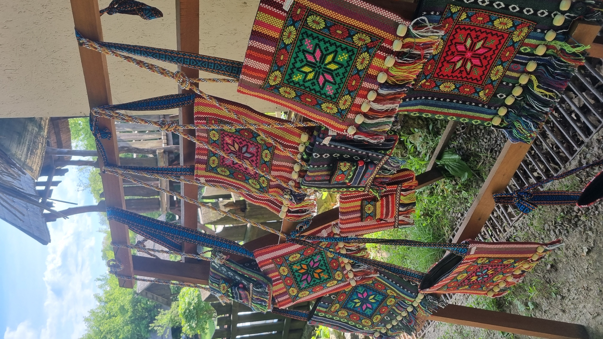 This image captures a static outdoor display of numerous handcrafted textile bags, reportedly located in the city of Buda, Ukraine. The bags are brightly colored, featuring intricate woven geometric patterns, primarily a central star or floral motif within a square, surrounded by vertical stripes. Predominant colors include red, green, blue, yellow, pink, and black. Many bags are adorned with multi-colored tassels and small spherical wooden beads along the bottom edge. Shoulder straps vary, with some being braided rope-like and others flat woven with complementary patterns. The bags are hung from a wooden display structure, consisting of horizontal bars and vertical supports made of light-colored timber. In the background, lush green trees and bushes are visible, along with a wooden fence and parts of a rustic wooden hut with a thatched or shingle roof. The sky is bright blue with scattered white clouds. The ground area visible includes dirt, gravel, and some green foliage, along with a metal grate. No individuals are present, and no actions or interactions are depicted.