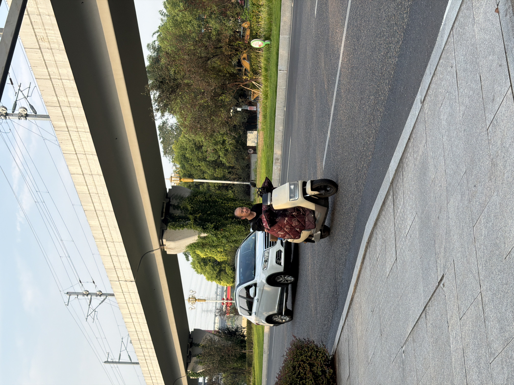 The image captures a street scene in Jiangjin, China, on a clear day. An elevated roadway or bridge structure, supported by concrete pillars, dominates the upper portion of the frame. Beneath the bridge, a white SUV is seen driving on the asphalt road, while a person on a beige scooter travels in the foreground. The scooter is covered with a patterned blanket. To the right of the road, a grassy area is visible with trees, a street lamp, and what appears to be construction equipment in the background. The sidewalk in the foreground is paved with gray tiles. The sky is light blue with some clouds.