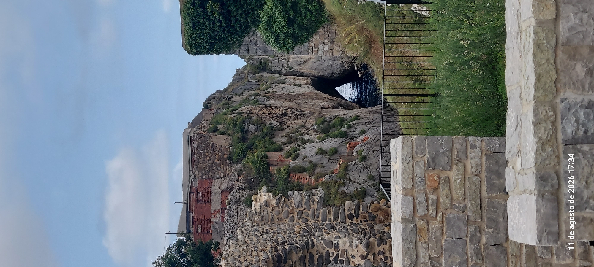 The image depicts a coastal scene in Castro-Urdiales, Spain, focusing on ancient stone structures and a natural sea cave. In the foreground, weathered stone walls and a low stone barrier create a sense of historical enclosure. Beyond this, the rugged coastline dominates, featuring a prominent cliff face with a dramatic sea cave opening. This cave reveals the dark blue water of the sea within.

The cliffs are partially covered with greenery and show remnants of brick structures, suggesting a past of construction or fortification integrated with the natural rock. A black metal fence runs horizontally across the middle ground, separating the viewer from the cliff face and sea. The sky above is a pale blue with scattered white clouds. The overall impression is one of weathered history and the raw beauty of the Cantabrian coast.