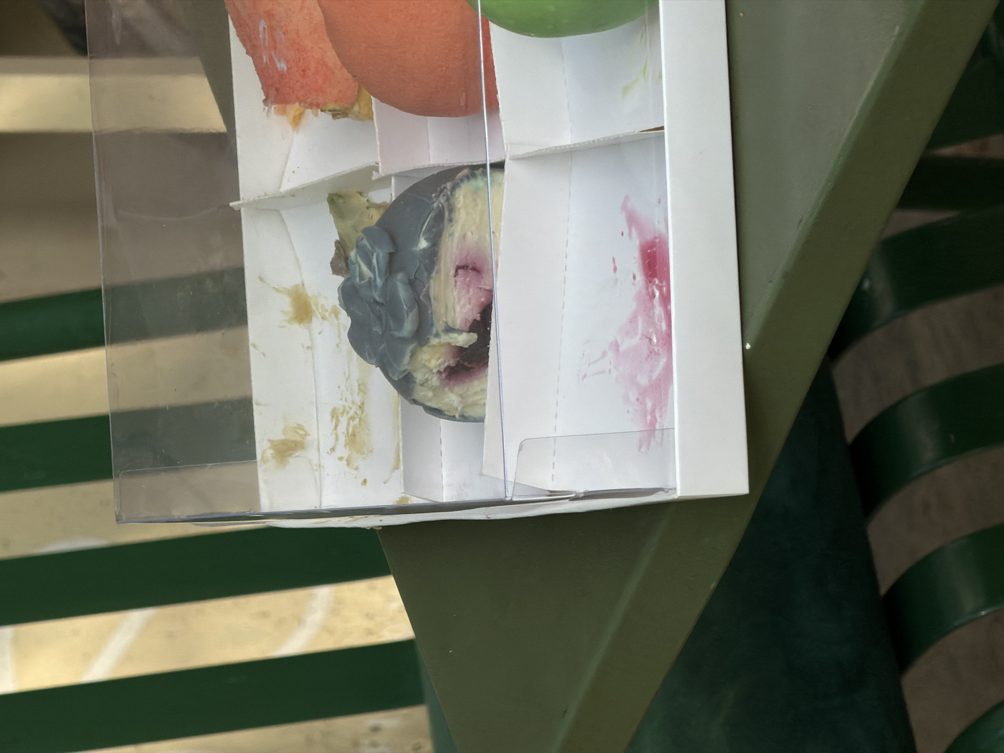 The image shows a clear plastic and white cardboard container with compartments, sitting on top of an olive-green surface that appears to be part of outdoor furniture, possibly a table or chair. The container holds decomposing food scraps or discarded items, including what seems to be moldy fruit. The background reveals a striped pattern in shades of green and off-white/beige, which may indicate a floor or wall. The items in the container appear messy and include discoloration or staining on the surrounding cardboard dividers. There is not enough information to identify the city, Bishkek City, Kyrgyzstan.