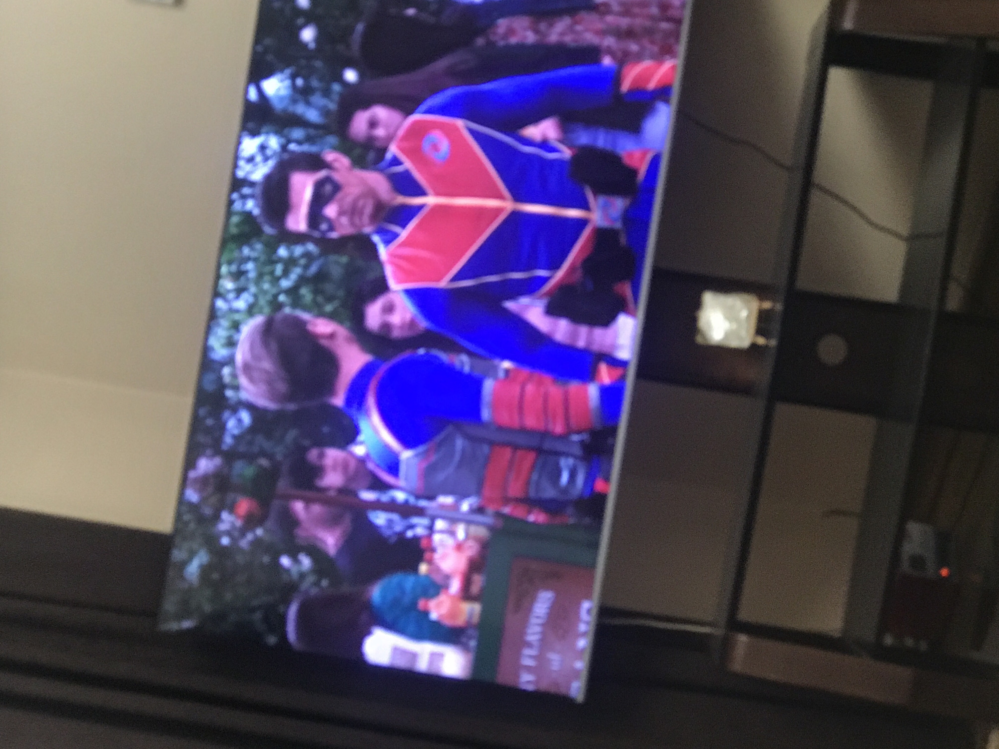 A television screen is displaying a scene from a TV show or movie featuring two individuals in superhero costumes. One superhero, facing the camera, wears a blue suit with red and yellow accents and a black eye mask. The second superhero, with blonde hair, wears a similar blue and red costume. A crowd of people is visible in the background. The television is placed on a dark wooden entertainment center. The location is unknown and cannot be determined from the provided image. It is not possible to tell if this is in Yerevan, Armenia.