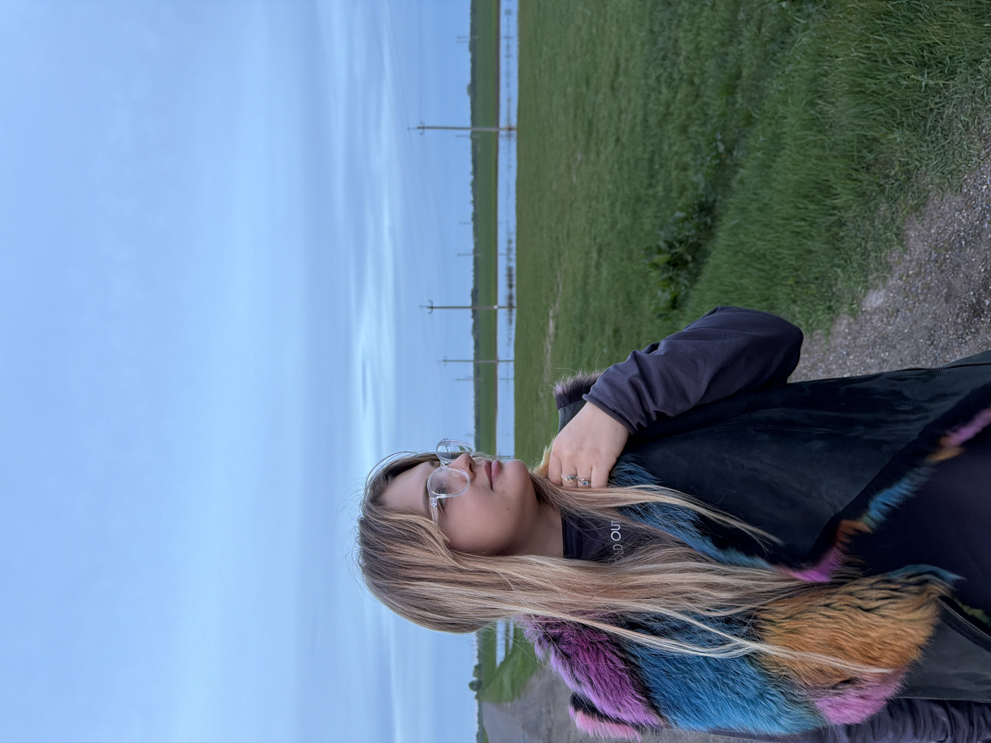 The image shows a woman with long blonde hair and glasses, standing outside in Azyakul', Russia. She is wearing a dark jacket with a colorful fur collar. In the background, there is a field of green grass, a flooded area, and power lines. The sky is overcast. The scene seems to be along a road or path on the edge of a field.