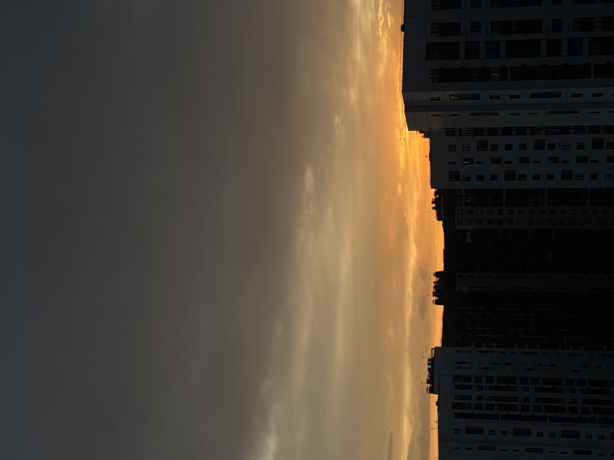 The image shows buildings silhouetted against a sunset in Lyubertsy, Russia. The buildings are multi-story residential structures with numerous windows. The sky transitions from a dark, overcast grey on the left to a bright, orange-yellow glow near the building outlines, indicating sunset. There are no visible people or specific objects besides the buildings and sky. The interaction is between the setting sun and the urban landscape. The environment is urban, during twilight.